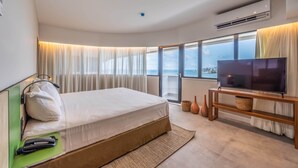 A modern hotel room with a large bed, a flat-screen TV on a wooden stand, a city view through large windows, and an air conditioning unit on the wall. - Novotel Salvador Rio Vermelho (Salvador)