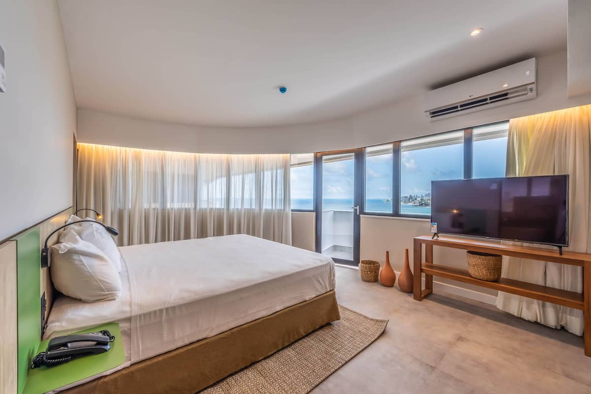 A modern hotel room with a large bed, a flat-screen TV on a wooden stand, a city view through large windows, and an air conditioning unit on the wall.