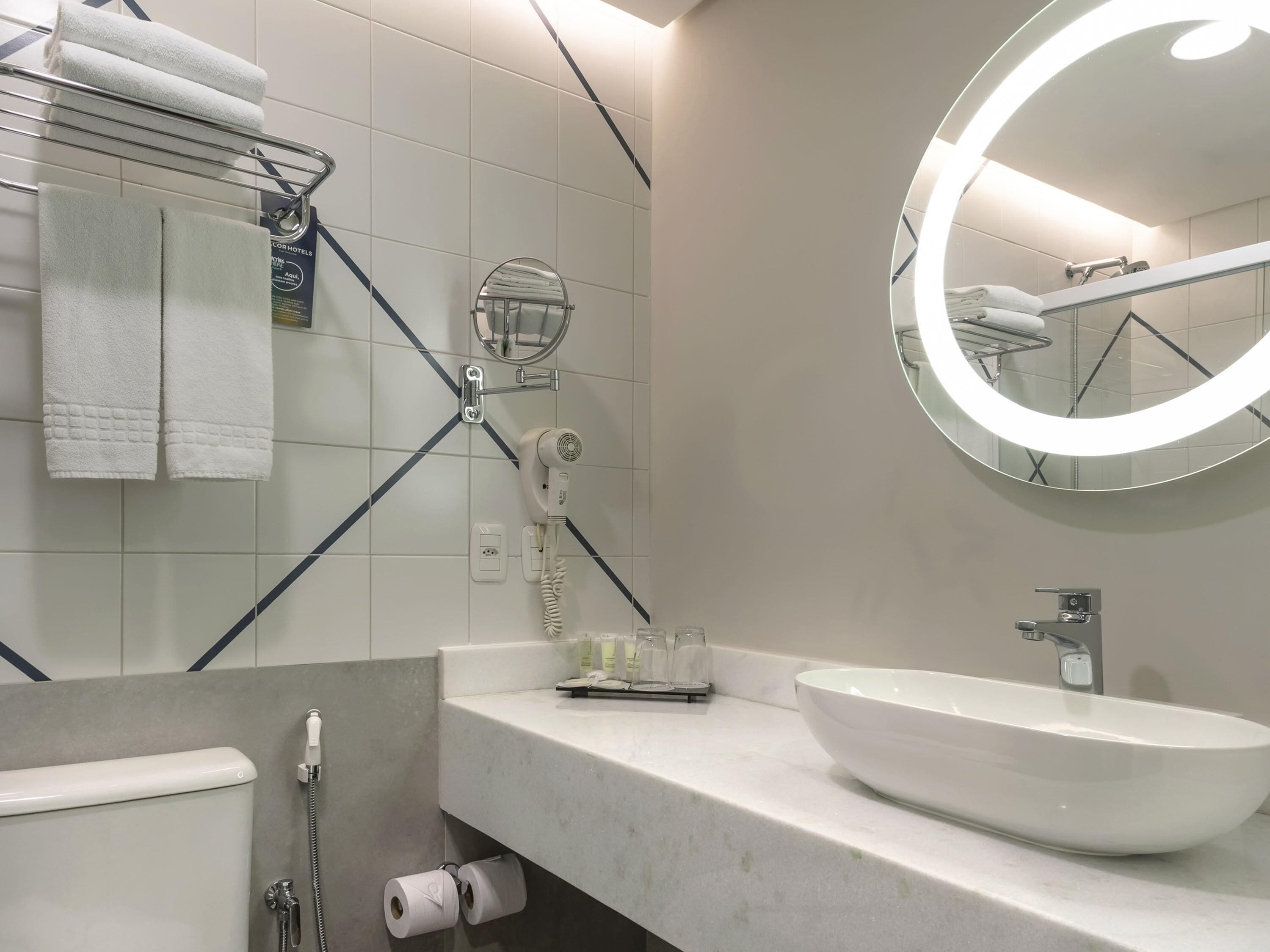 A modern bathroom with a white sink, a round mirror with a light, a towel rack, and a hairdryer.