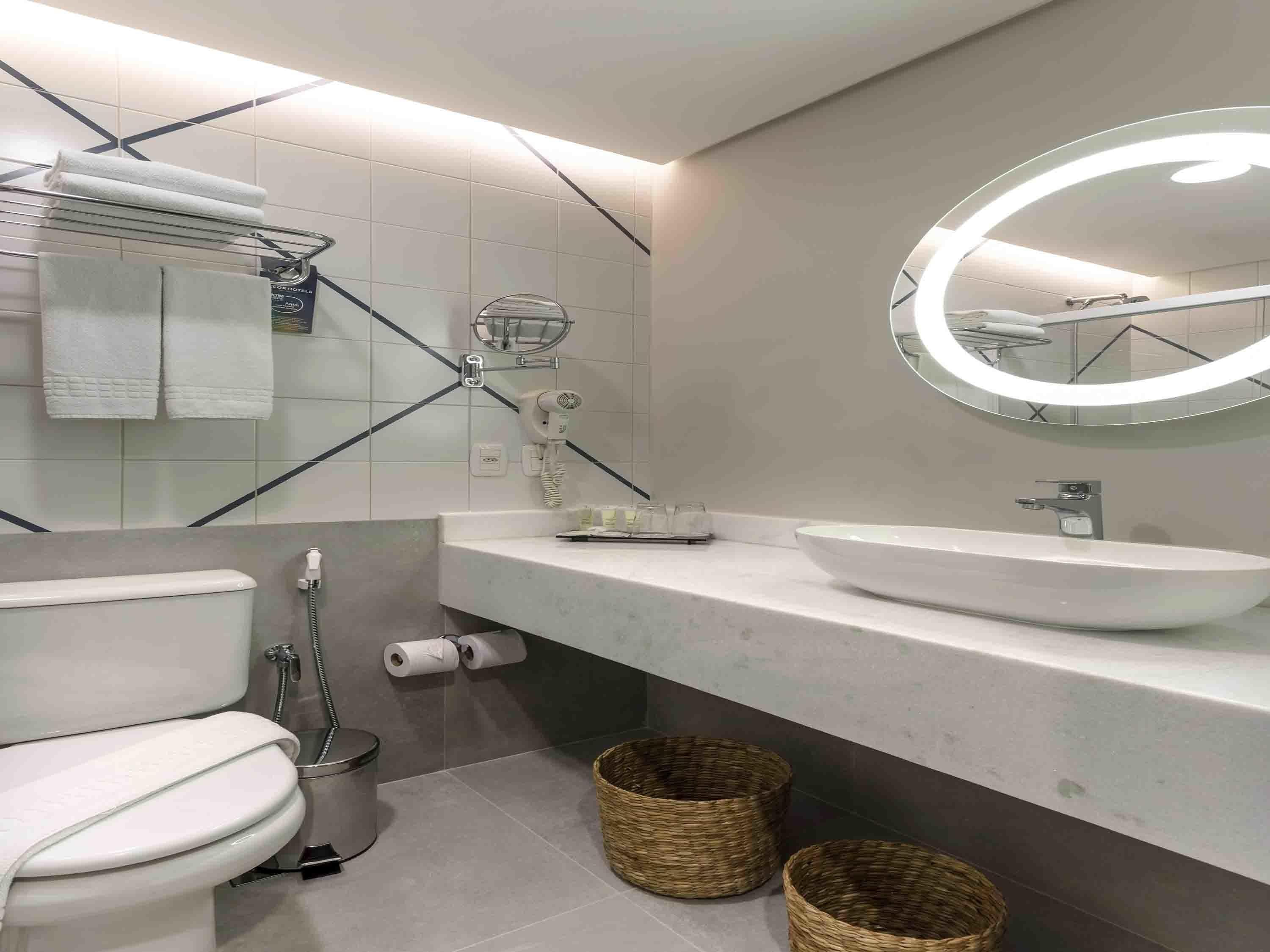 Un baño moderno con inodoro, bidé, lavabo y un espejo con luz circular.