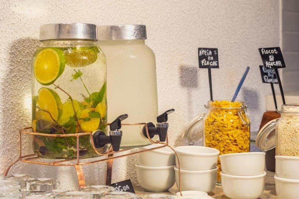 Un dispensador de bebidas con una rodaja de lima y menta, junto a un recipiente grande y frascos con alimentos.