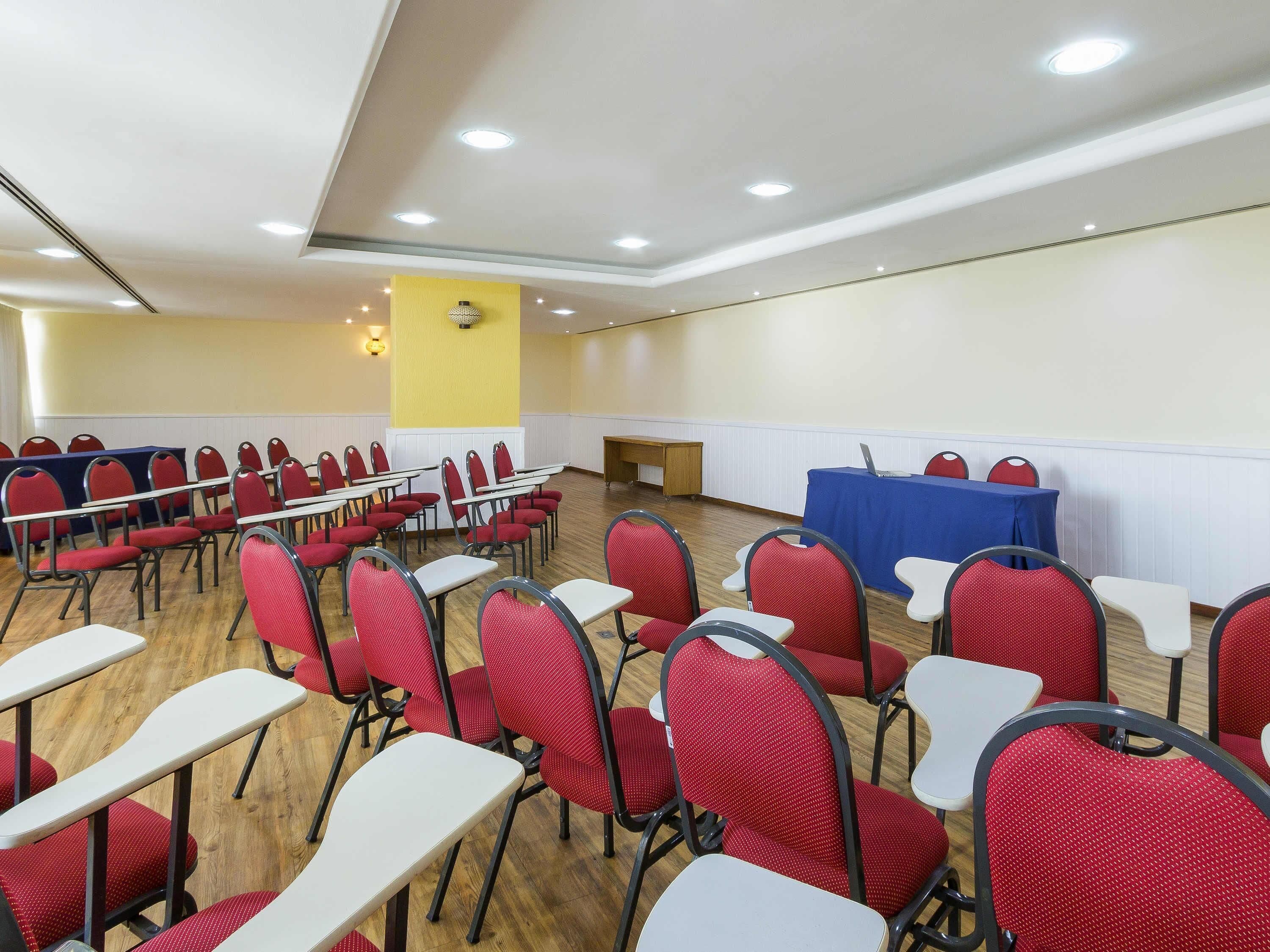 Una sala de conferències amb files de cadires vermelles i taules blanques, equipada amb llums al sostre.