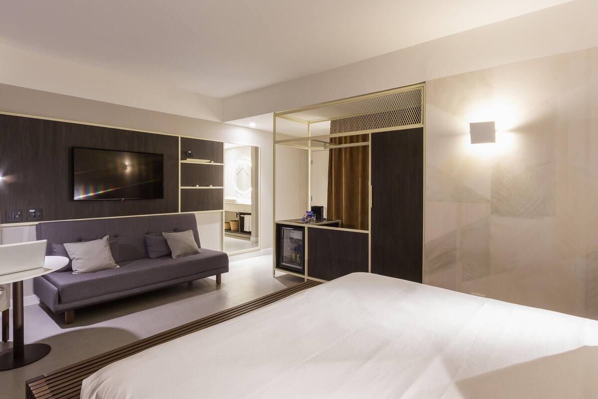 A modern hotel room with a large bed, a sofa, a TV, and a mirror.