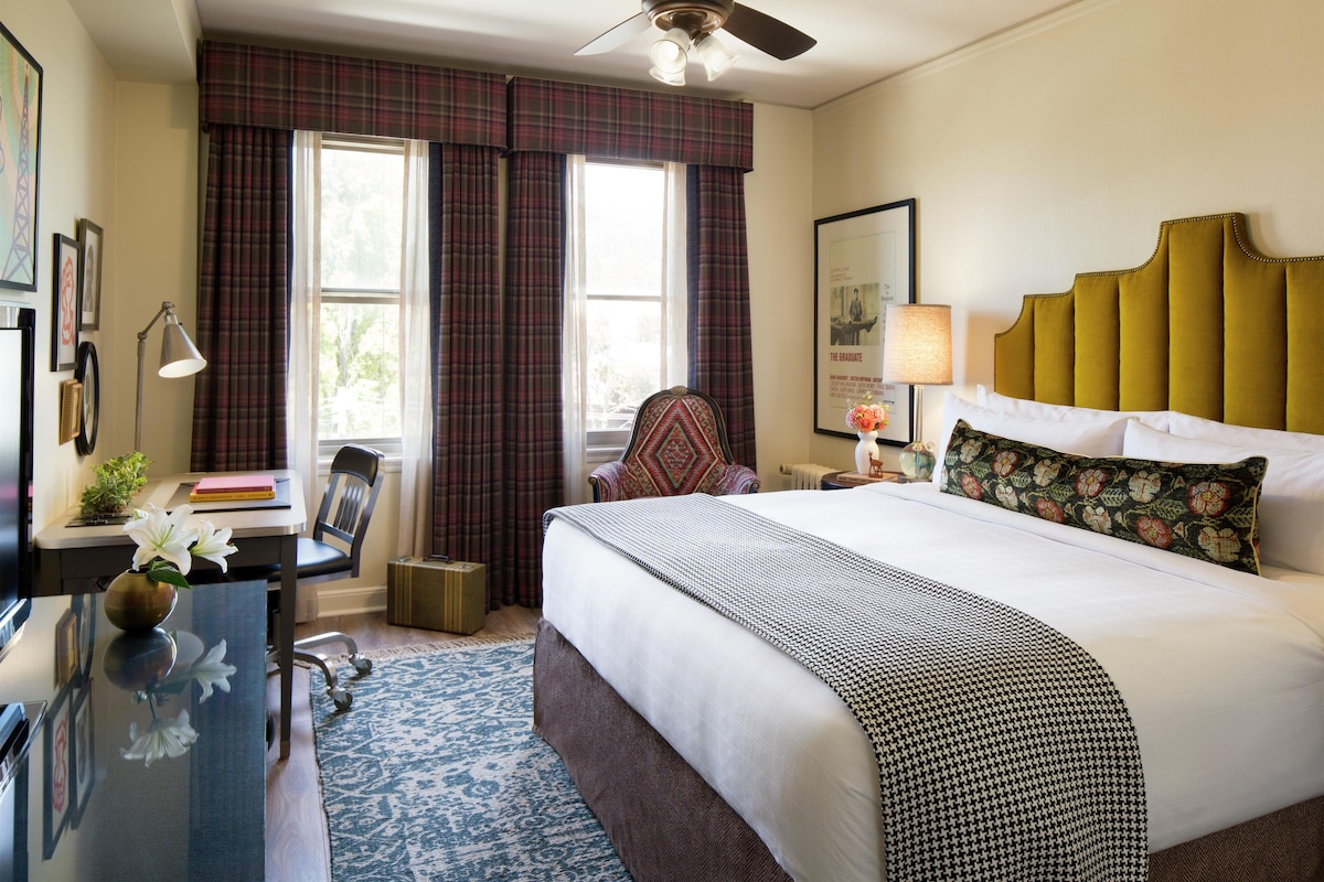 A hotel room with a bed, a desk with a lamp, a chair, a desk lamp, a ceiling fan, and a window with curtains.