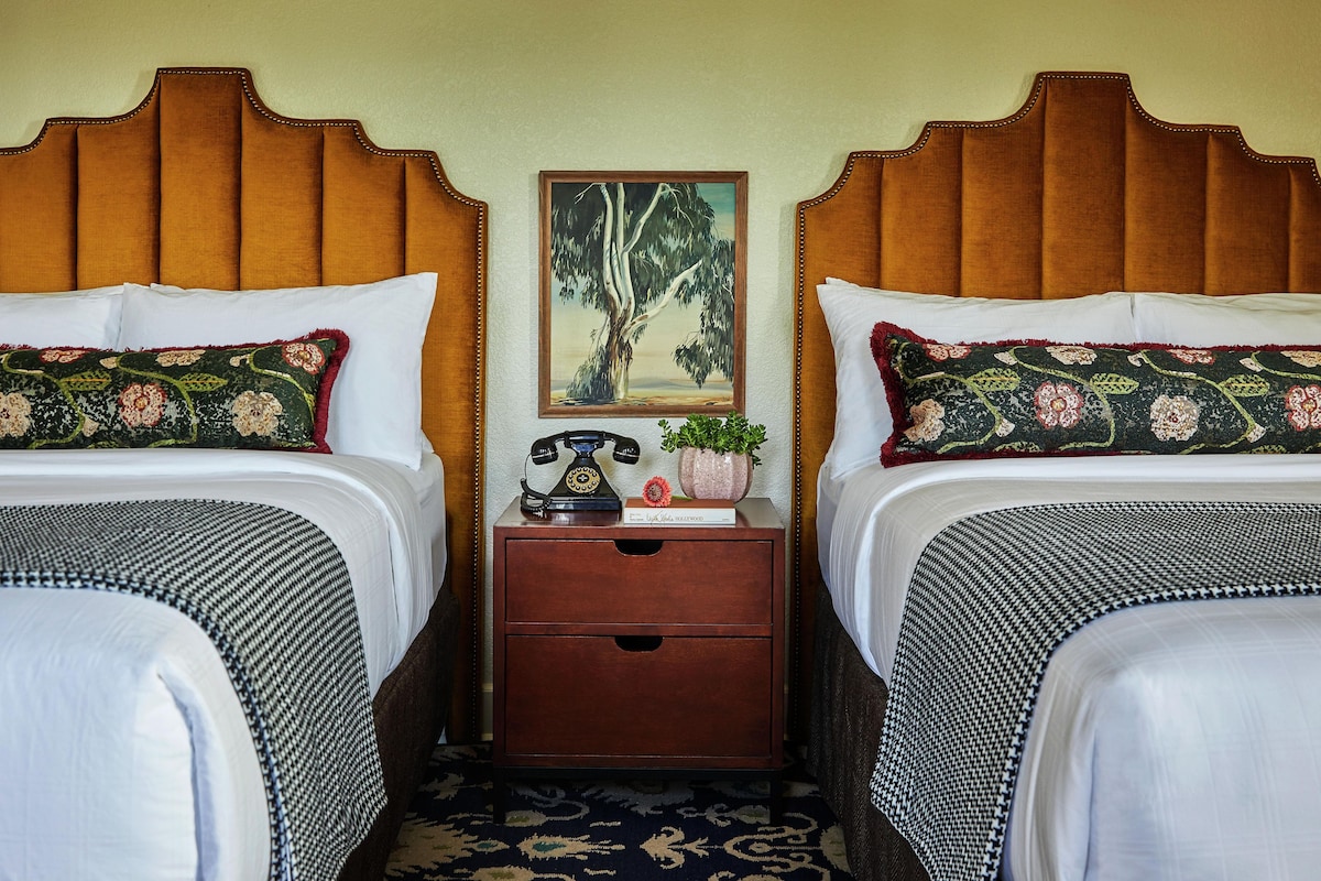 A hotel room with two beds, a nightstand with a telephone, a framed painting, and a potted plant.
