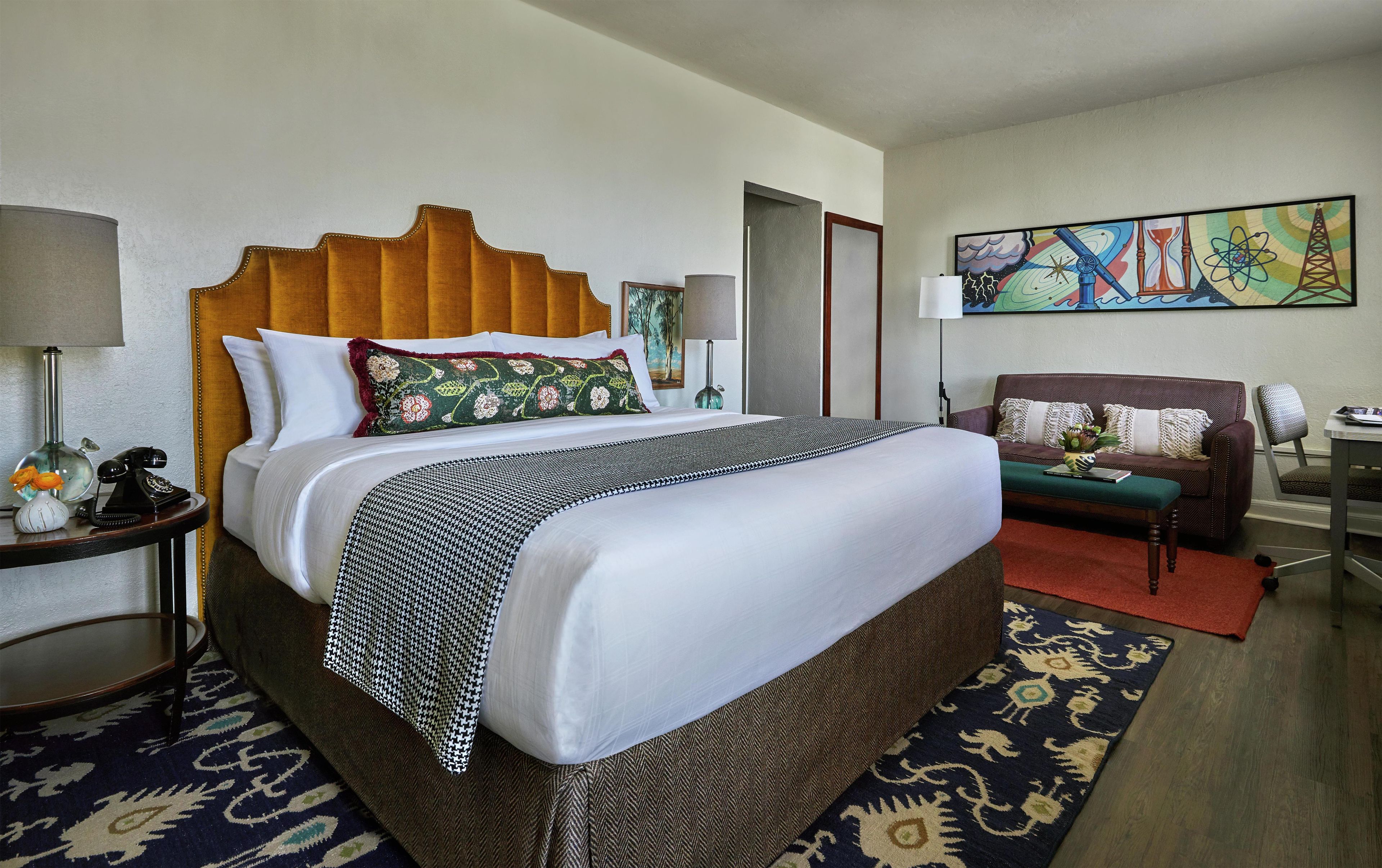 Quarto de hotel com uma cama grande, um sofá, uma poltrona, um abajur, um quadro e uma mesinha.