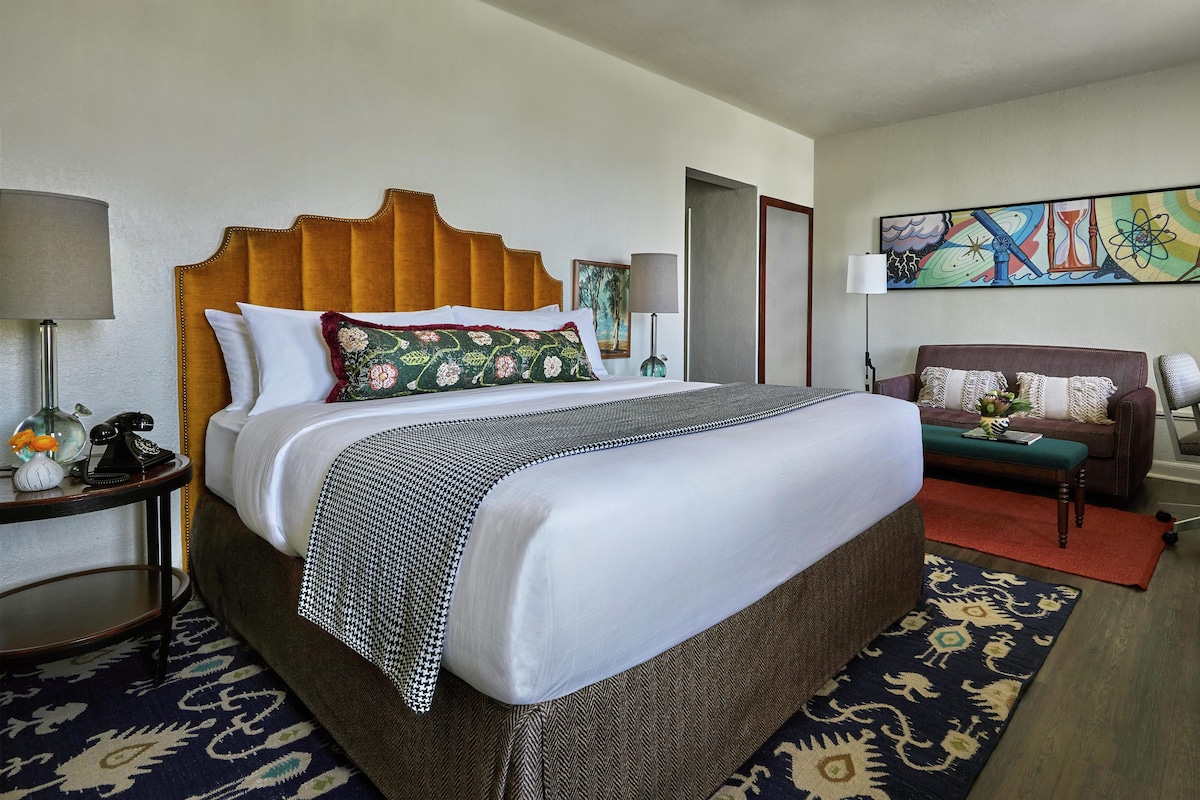 A hotel room with a large bed, a sofa, a chair, a lamp, a painting, and a small table.