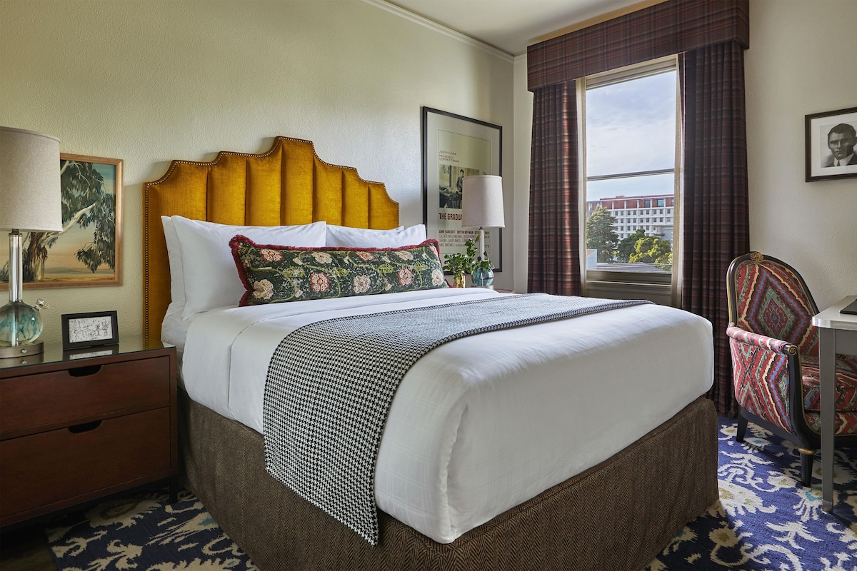 A hotel room with a large bed, a desk, a chair, a window with curtains, and a framed picture on the wall.
