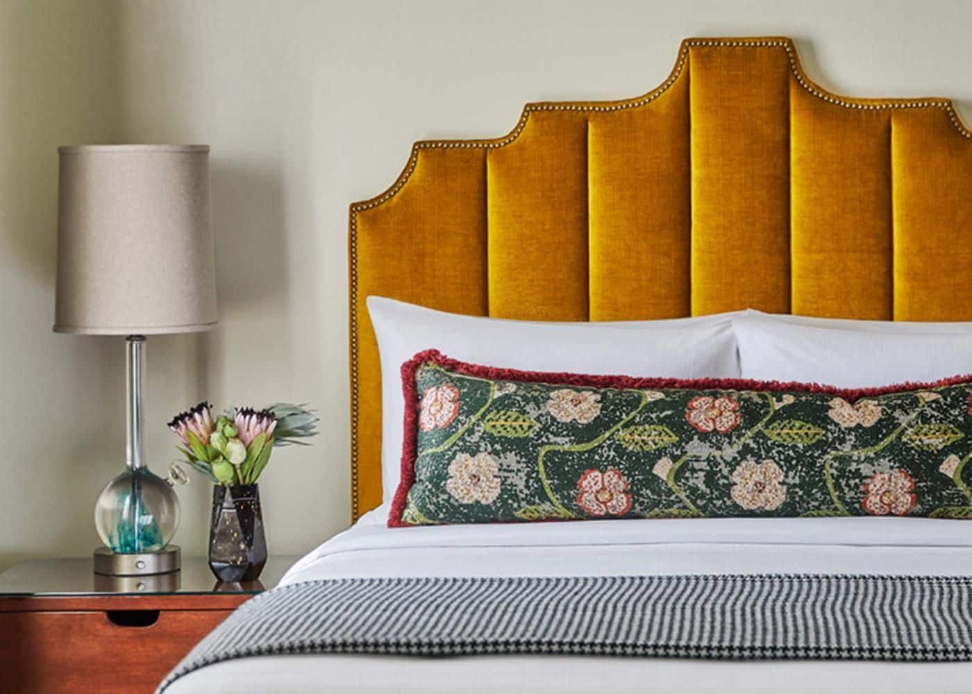 Una cama con cabecera capitonada, una lámpara de noche, un florero con flores y un cojín decorativo.