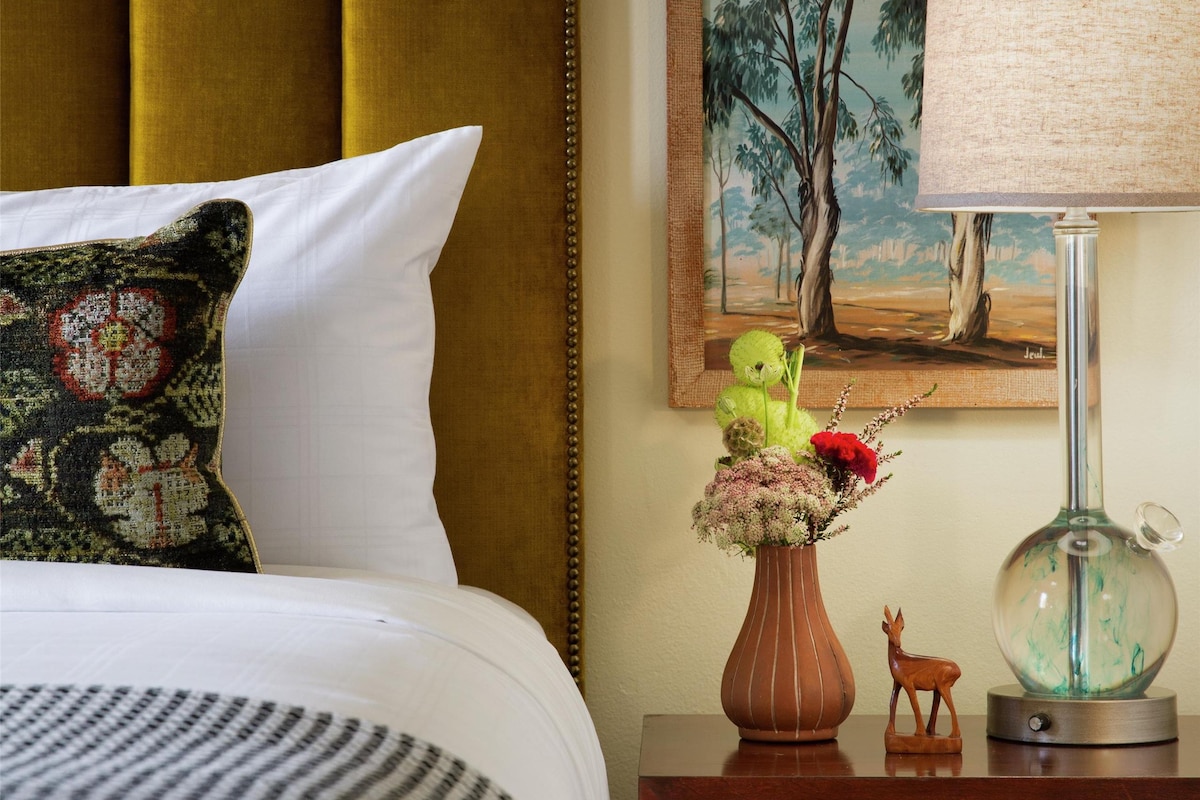 A bed with a headboard, a bedside table with a lamp, a vase with flowers, and a painting on the wall.