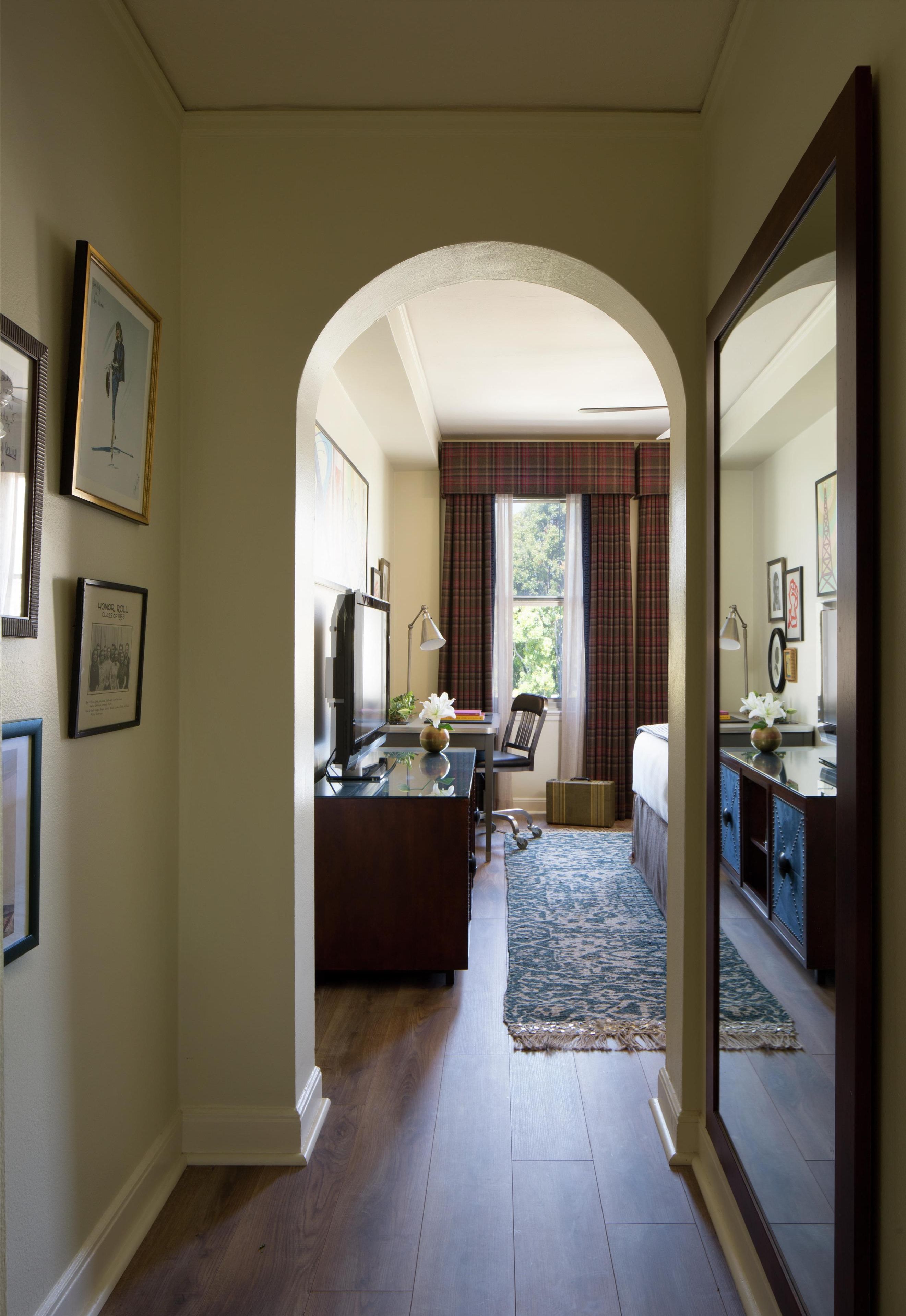 A hallway leading to a bedroom with a bed, a desk with a chair, a television, and framed pictures on the walls.