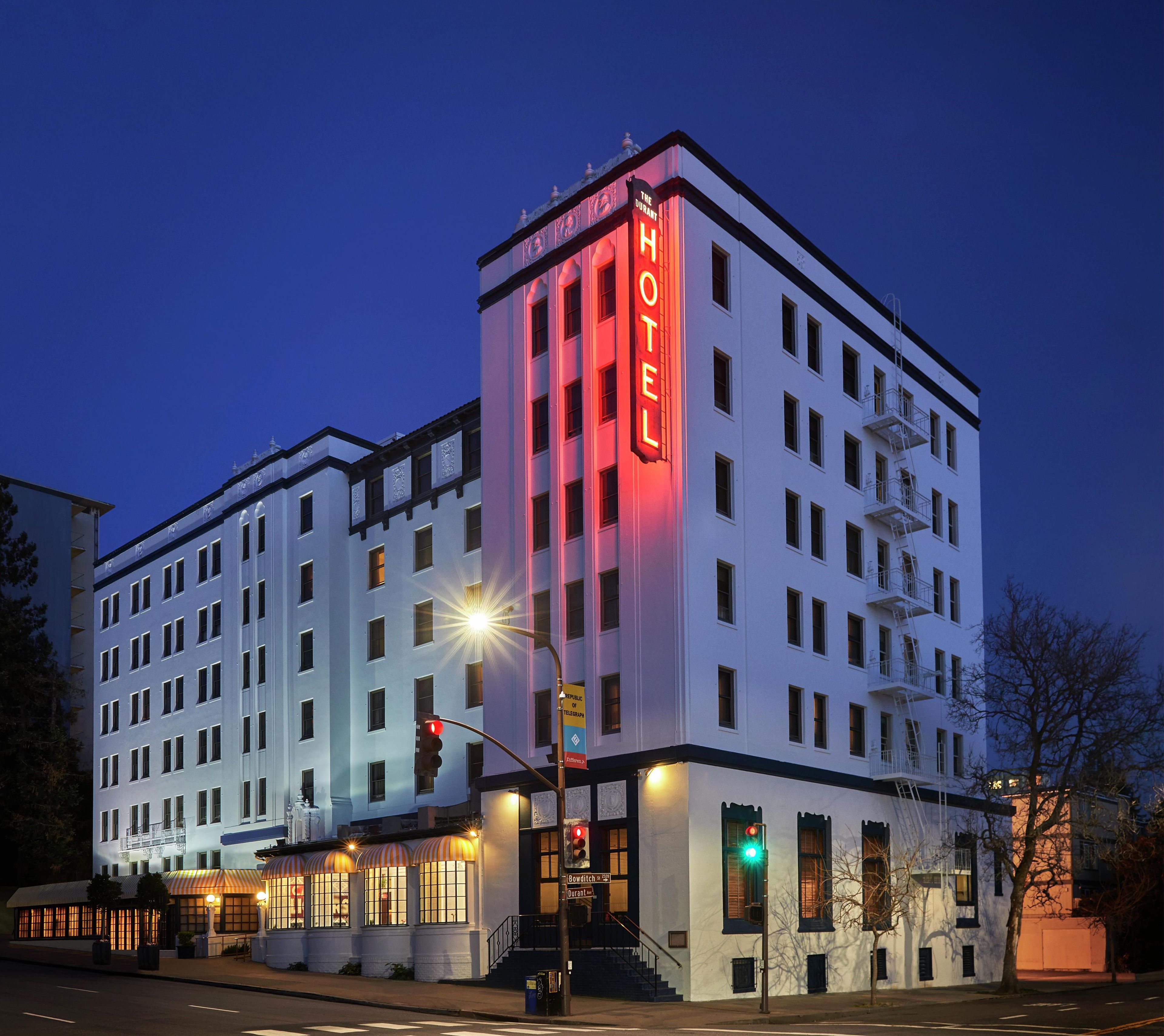 Un hotel de varios pisos con un llamativo letrero que dice 'HOTEL' en la esquina, una entrada visible con escalones, y una vista de la calle con semáforos.