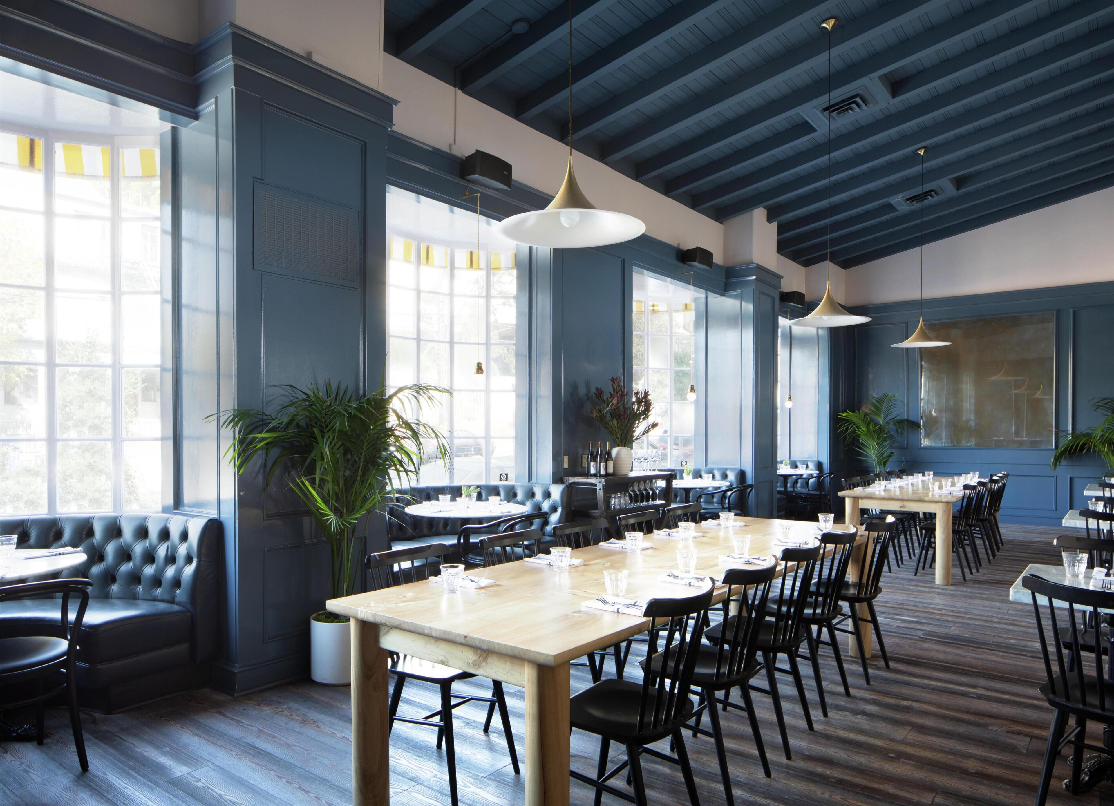 A restaurant interior with high ceilings, large windows, leather seating, and wooden tables.