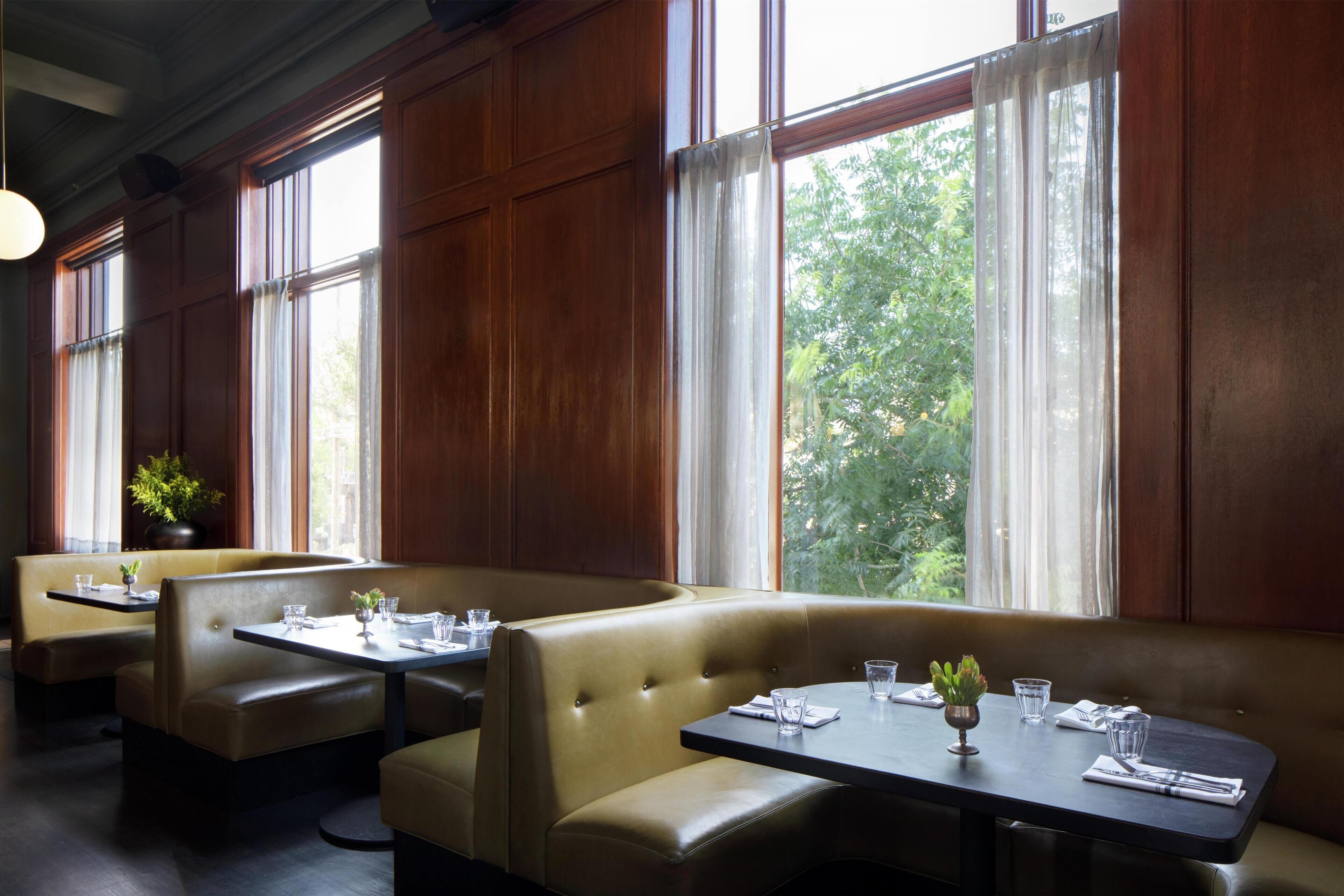 A restaurant with booths and tables, large windows with curtains, and a plant on a table.