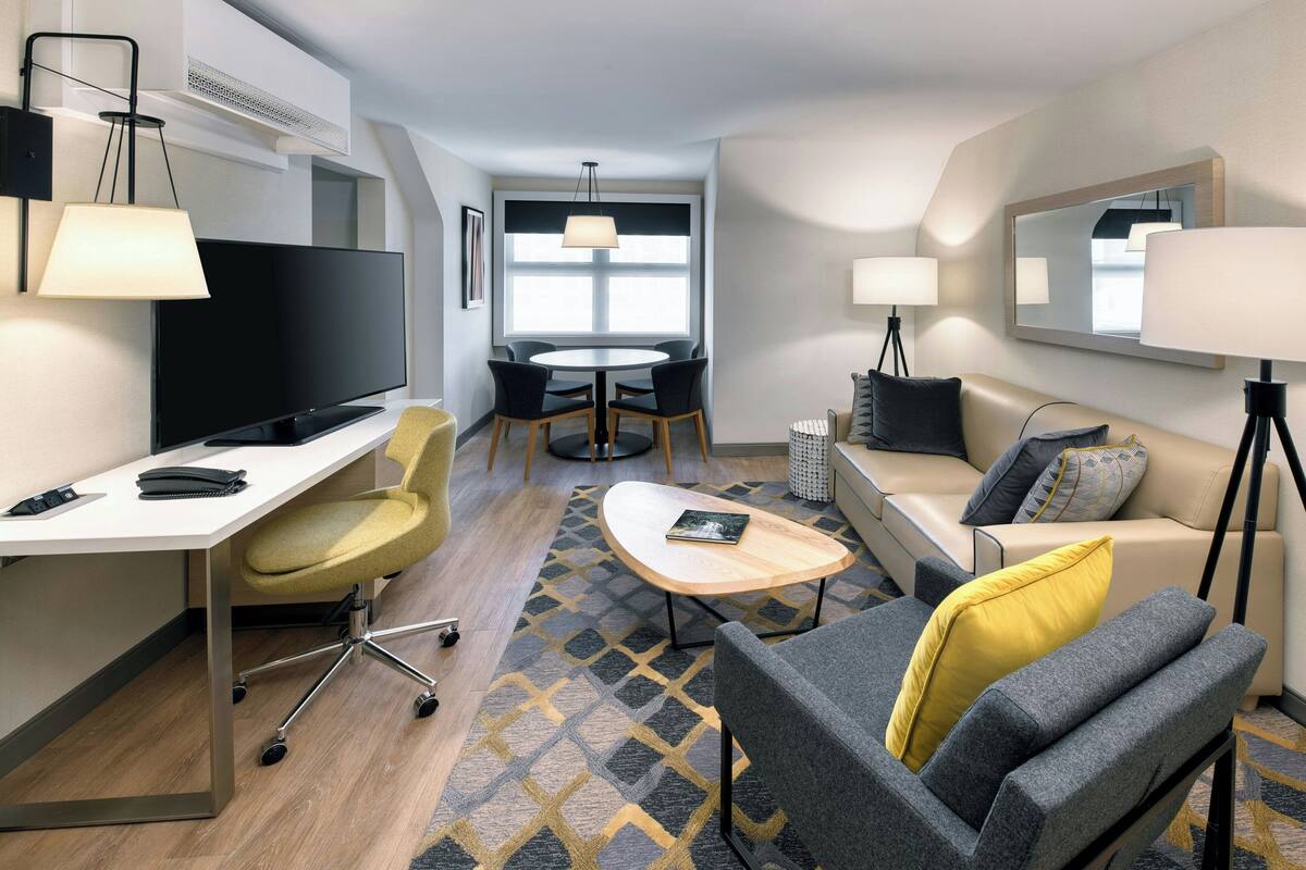 A modern hotel room with a flat-screen TV, a desk with a chair, a sofa, a coffee table, and a dining area with chairs and a table.
