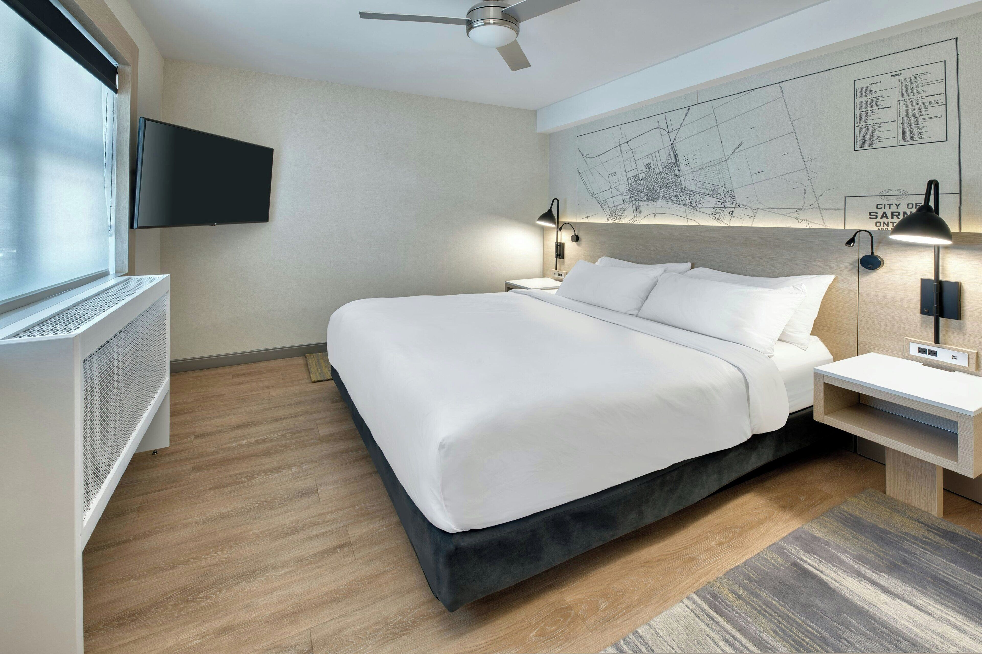 A modern hotel room with a large bed, a flat-screen TV mounted on the wall, a ceiling fan, and a map of the city on the wall.