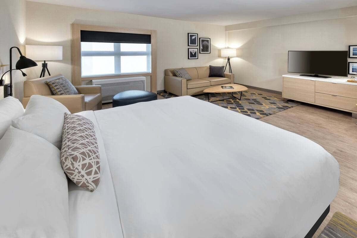 A hotel room with a large bed, a sofa, a small table, a TV, and a window with blinds.