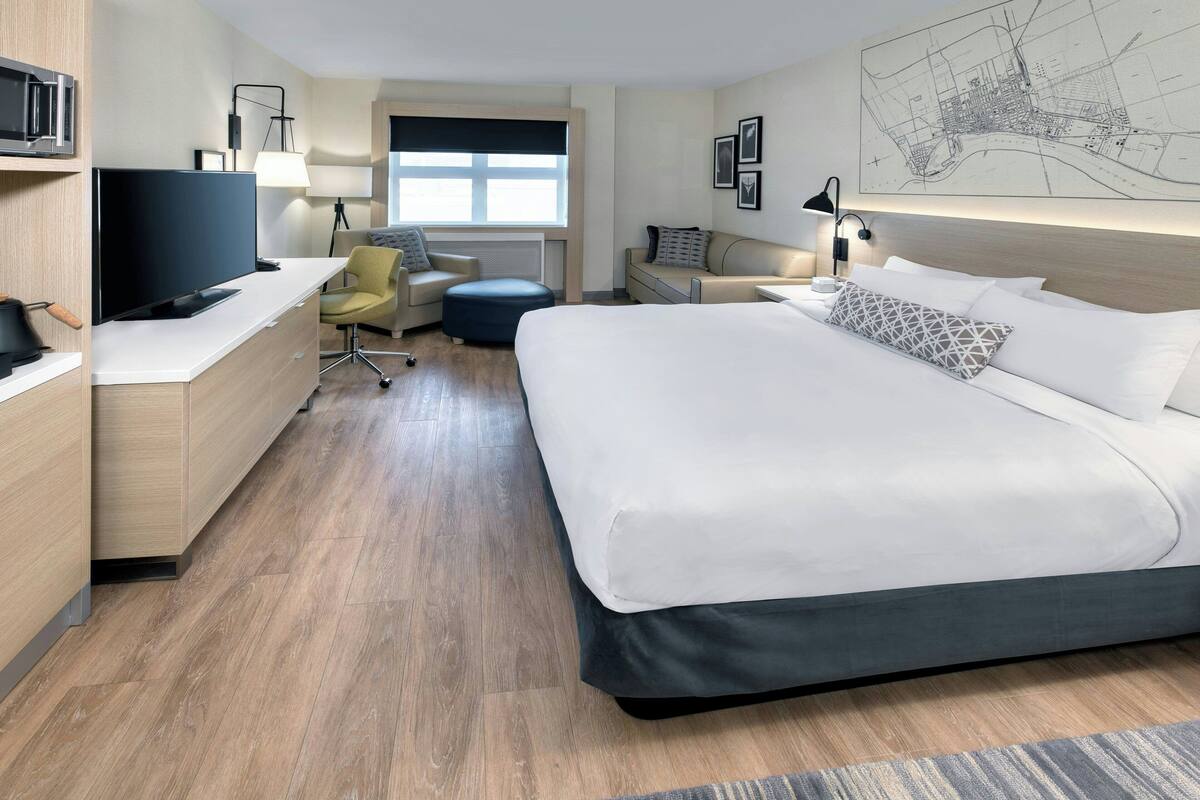 A modern hotel room with a large bed, a desk with a flat-screen TV, a chair, a sofa, and a map on the wall.
