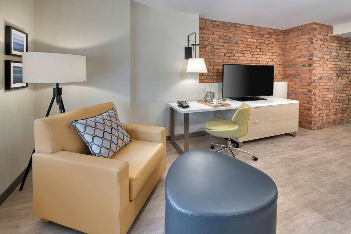 A modern hotel room with a beige sofa, a blue ottoman, a desk with a computer and a flat-screen TV, a lamp, and a brick accent wall.