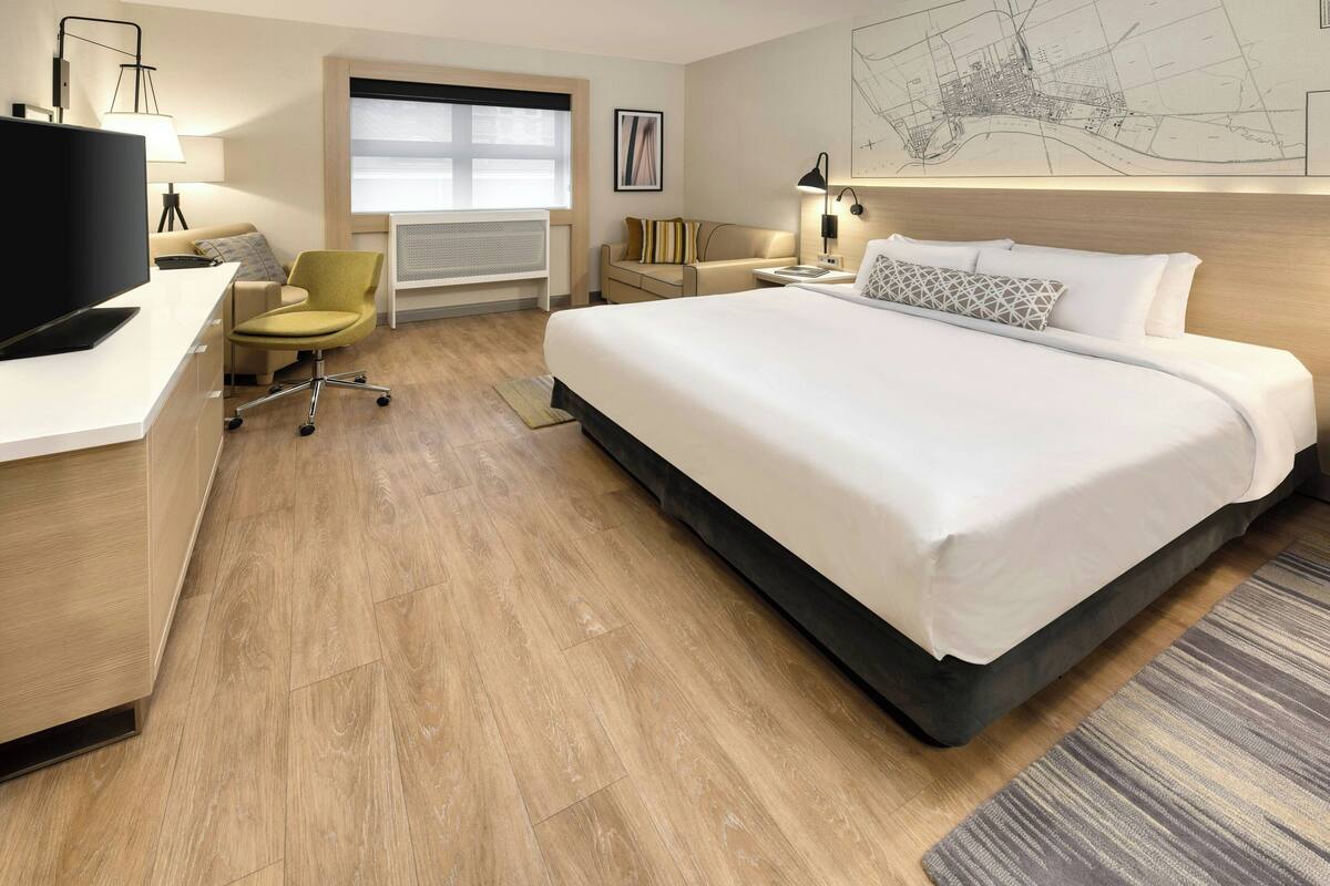 A hotel room with a large bed, a desk with a computer, a chair, a window with blinds, a map on the wall, and a radiator.