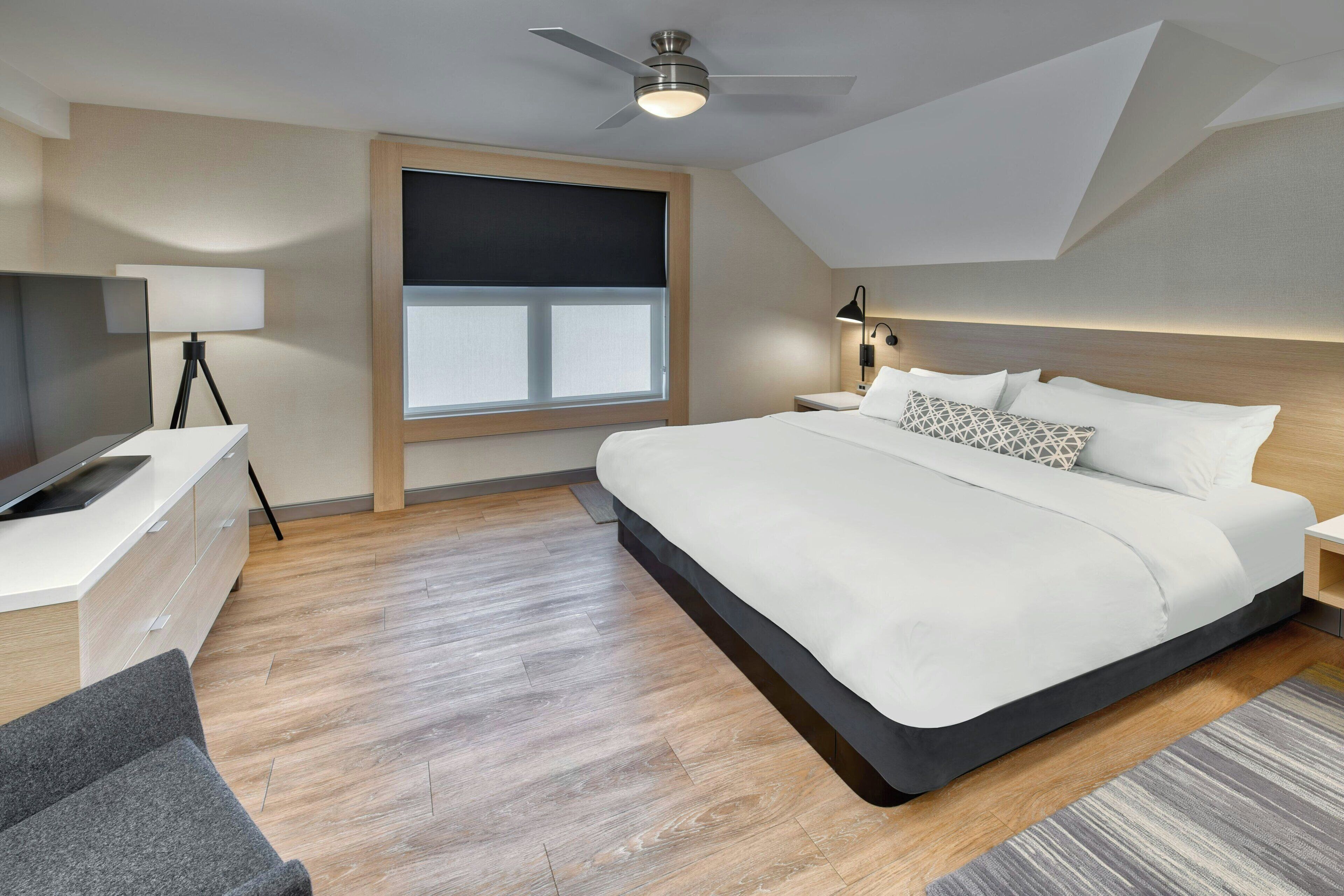 A modern hotel room with a large bed, a desk with a computer, a ceiling fan, and a large window with blinds.