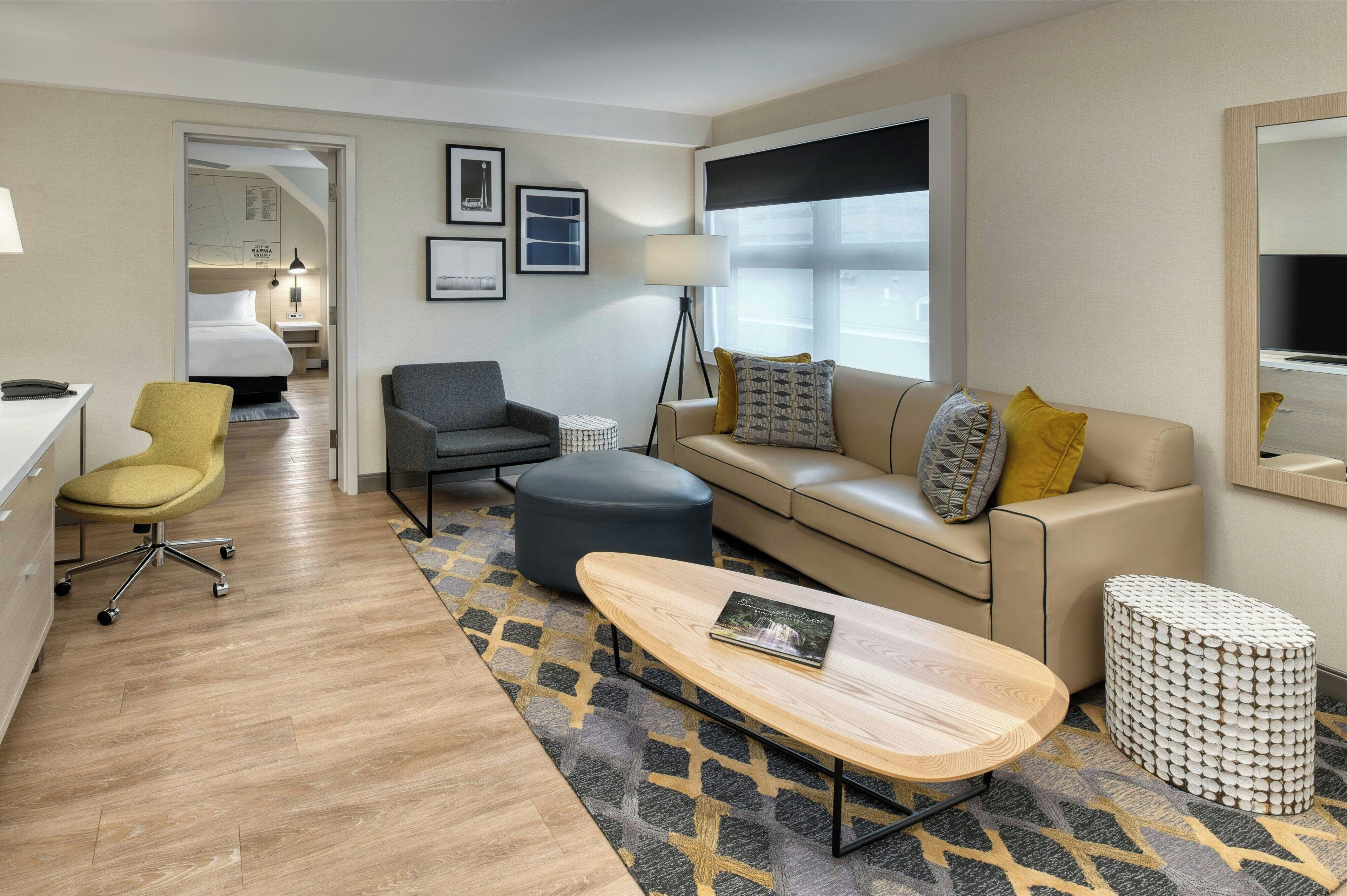 A modern hotel room with a sofa, armchair, ottoman, and a coffee table. There is a bed in the background, a lamp, and a framed picture on the wall.