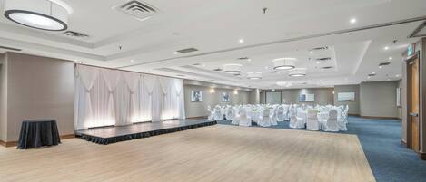 Un salón amplio con un escenario, filas de mesas con manteles blancos y una alfombra azul.