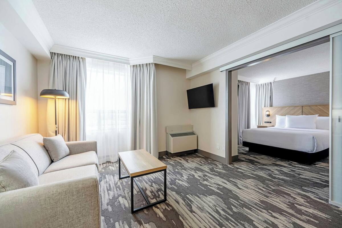 A hotel room with a sofa, a small table, a bed, a TV, and a sliding door leading to another room.