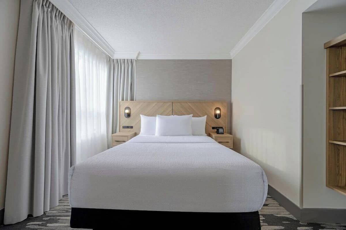 A hotel room with a large bed, two bedside tables, a headboard with integrated lighting, and a large window with curtains.