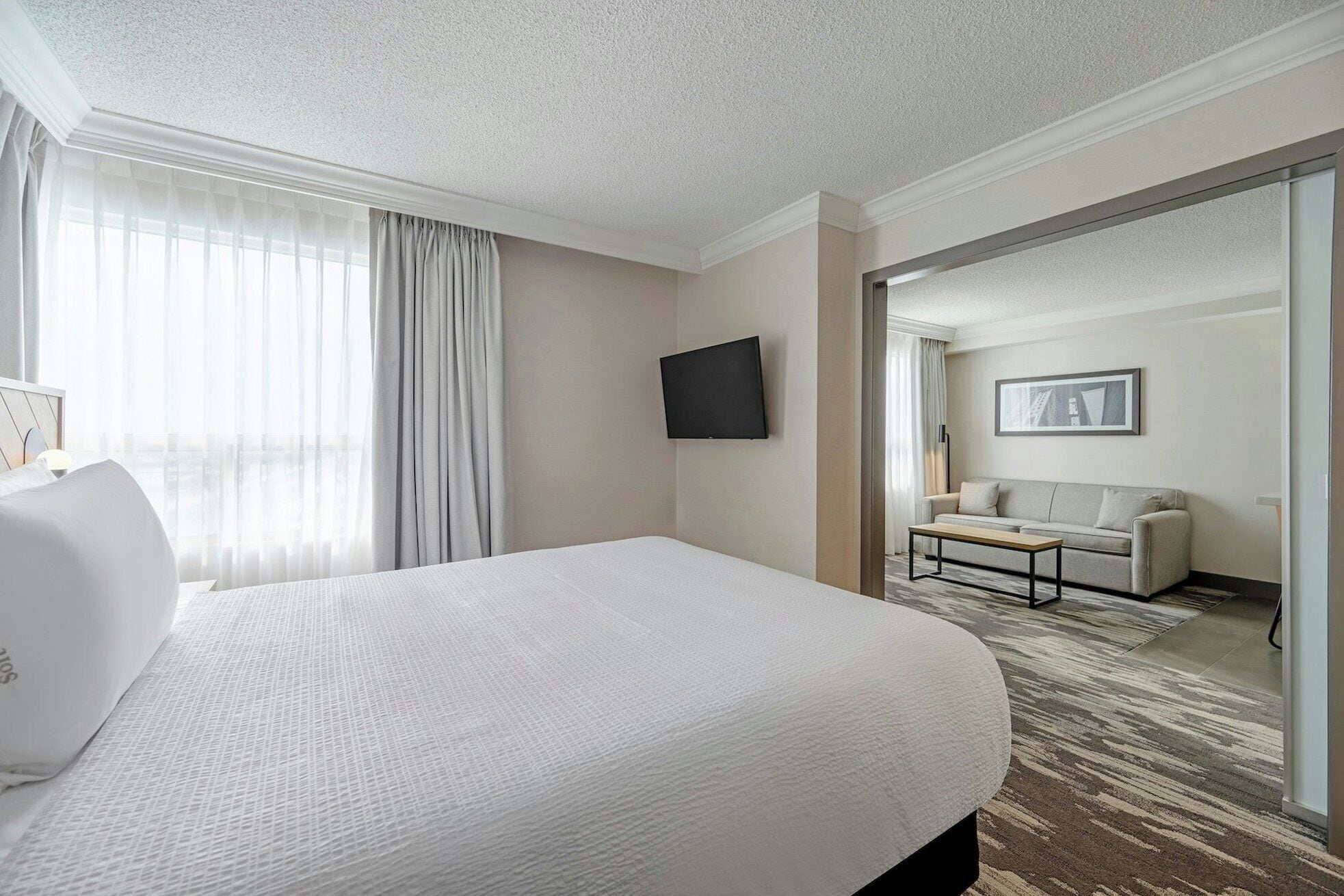 A hotel room with a large bed, a flat-screen TV mounted on the wall, a sofa, and a door leading to another room.