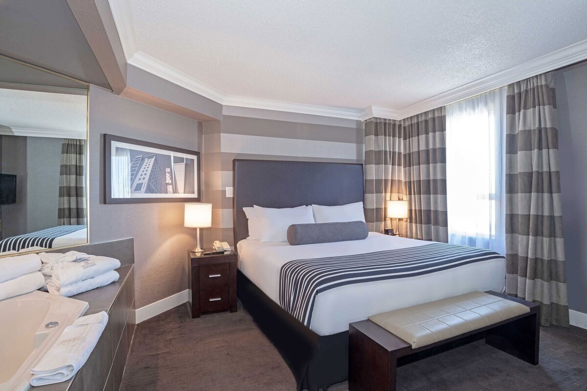 A hotel room with a large bed, bedside lamps, a bench, a bedside table with a phone, a mirror, and a framed picture on the wall.