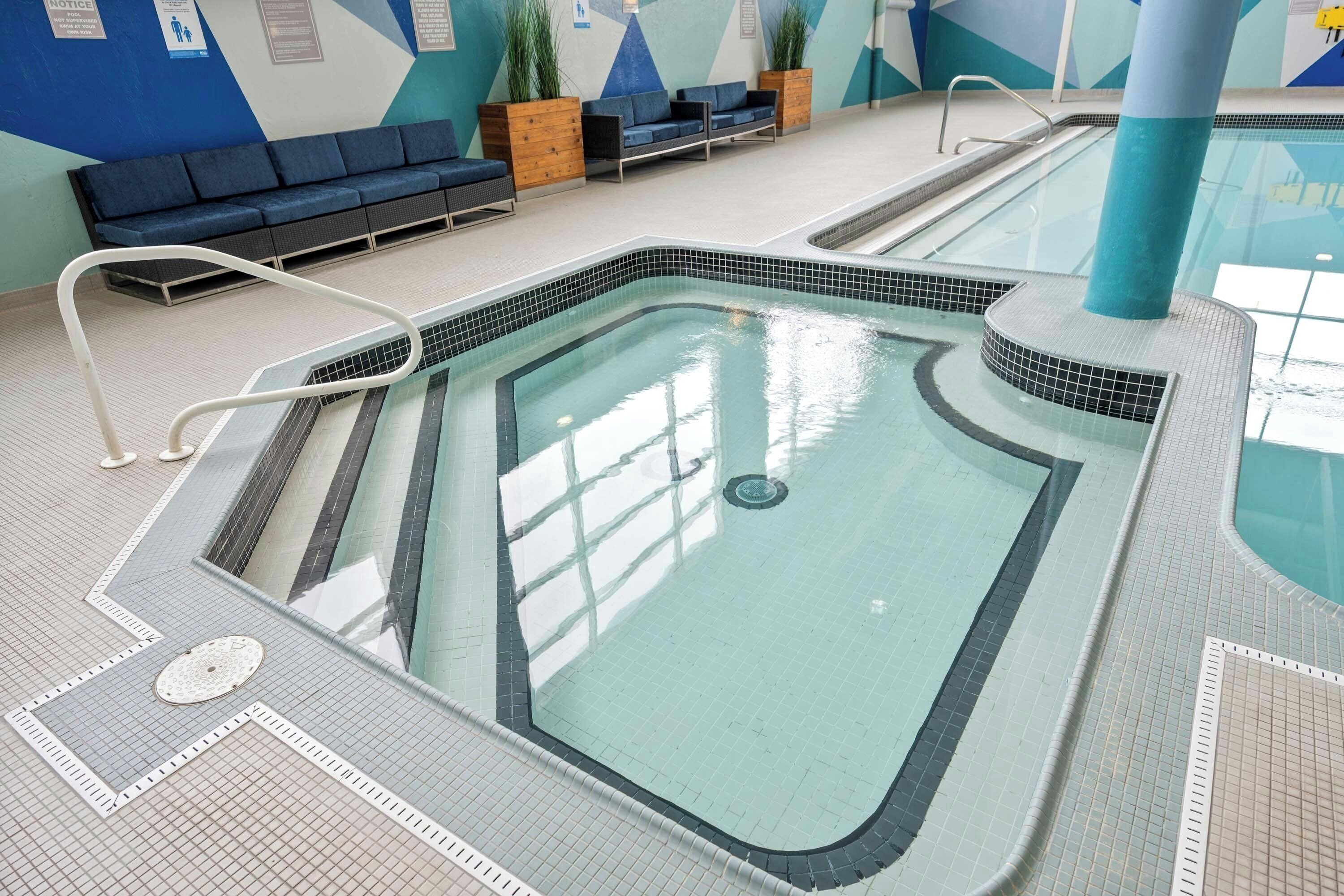 An indoor swimming pool with a tiled floor, a seating area with blue chairs, and a wall with informational posters.
