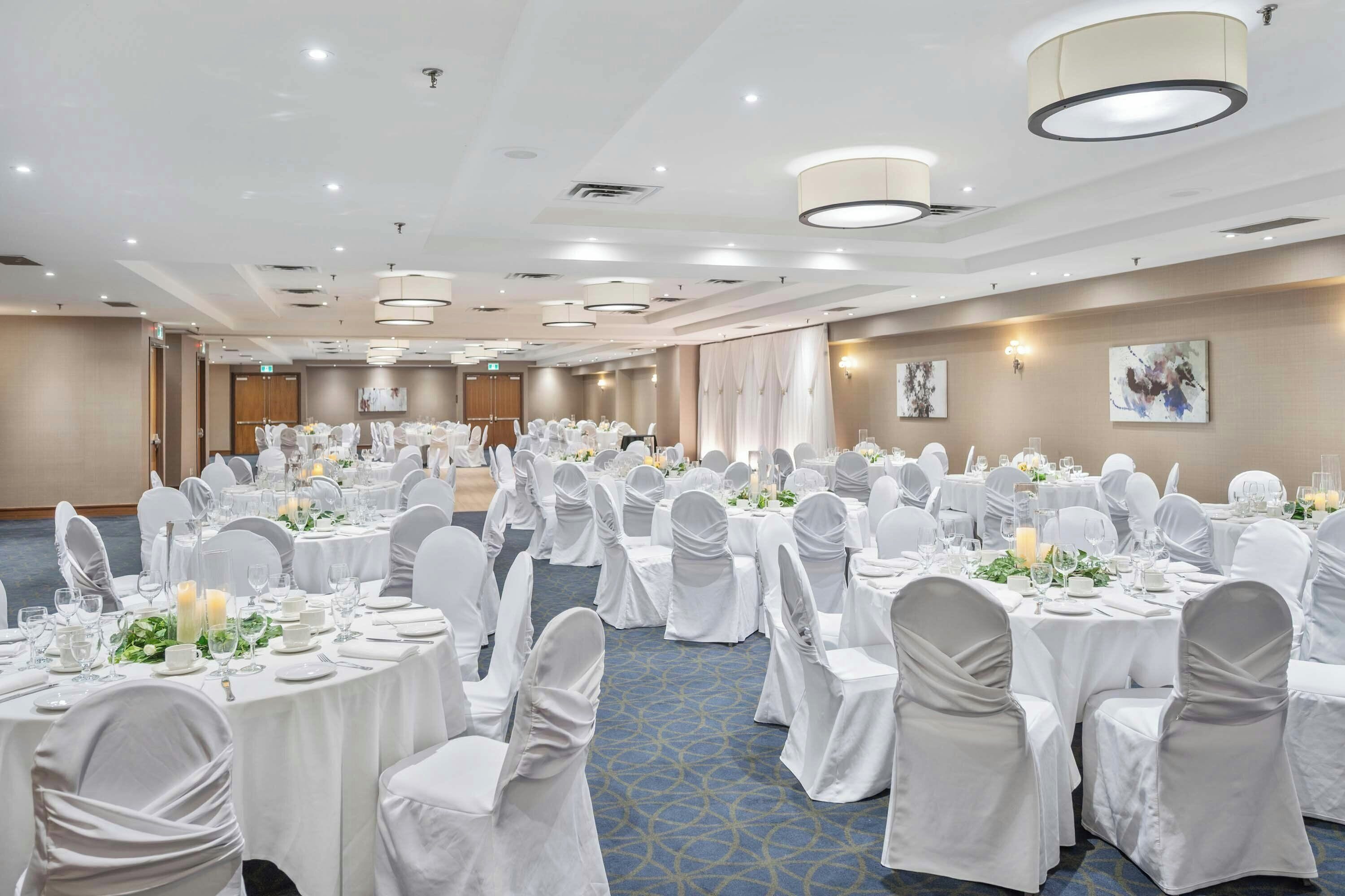 A large banquet hall with round tables set for a formal event, featuring white tablecloths and chairs, and a blue carpeted floor.