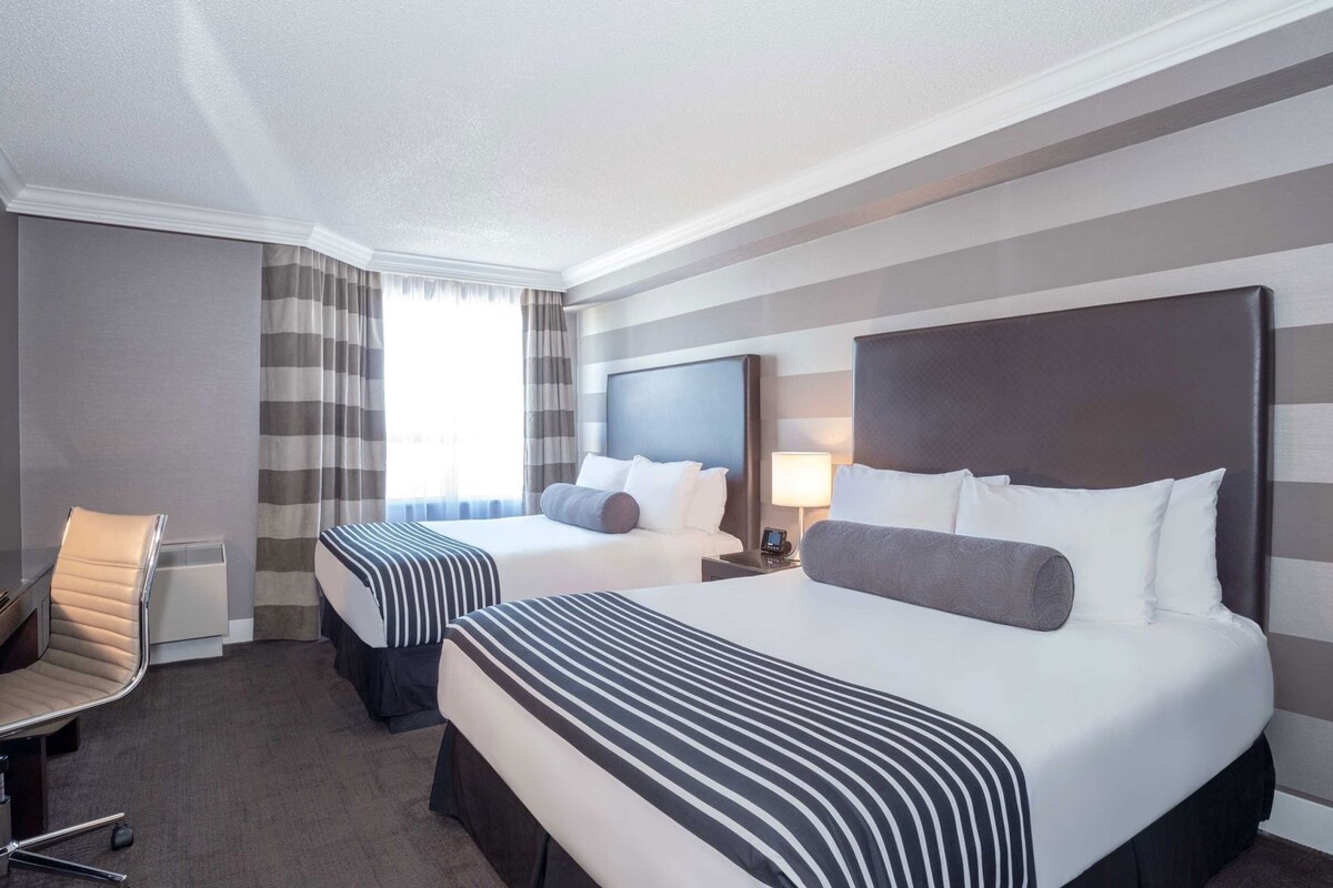 A hotel room with two beds, a chair, a desk, and a large window with curtains.