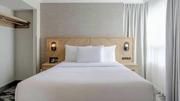 A hotel room with a large bed, a wooden headboard, two bedside tables with lamps, and a view of the outside through sheer curtains.