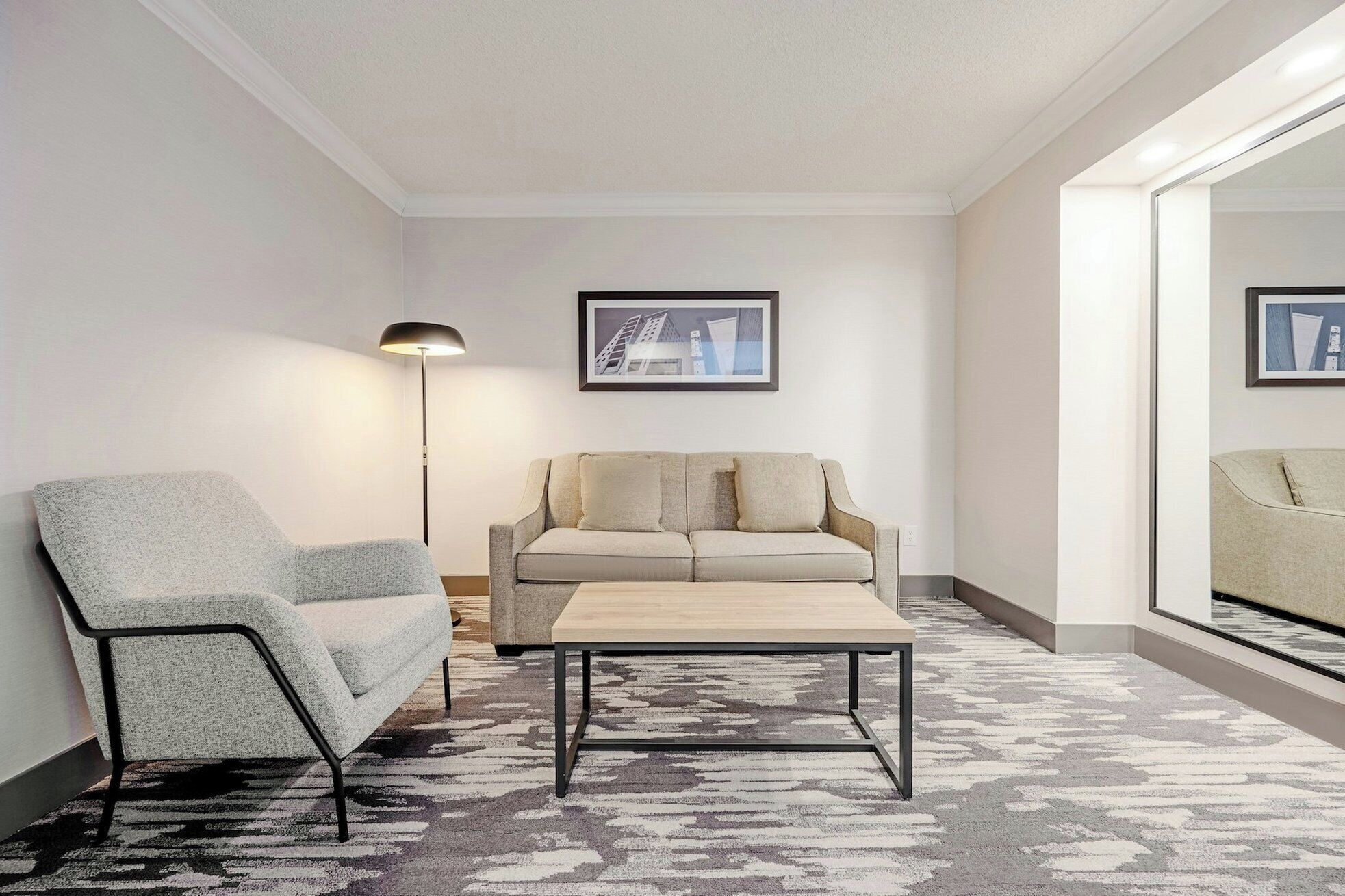 En moderne stue med en grå lænestol, en beige sofa og et lille træsofabord. Der er en gulvlampe og et indrammet billede på væggen.