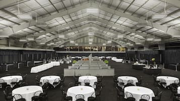 A large indoor event space with multiple round tables and chairs, a stage, and a high ceiling with industrial lighting.