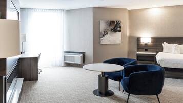 Una habitación de hotel moderna con una cama, un escritorio, dos sillas, una mesa redonda, un cuadro en la pared y una mesita de noche con una lámpara.