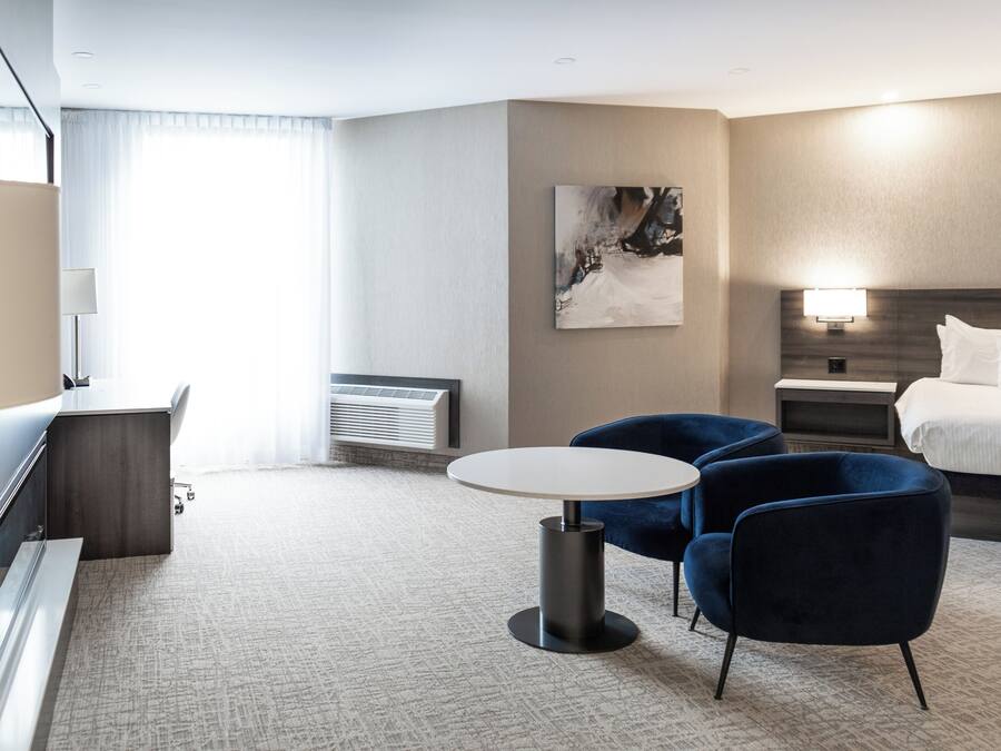 Una habitación de hotel moderna con una cama, un escritorio, dos sillas, una mesa redonda, un cuadro en la pared y una mesita de noche con una lámpara.