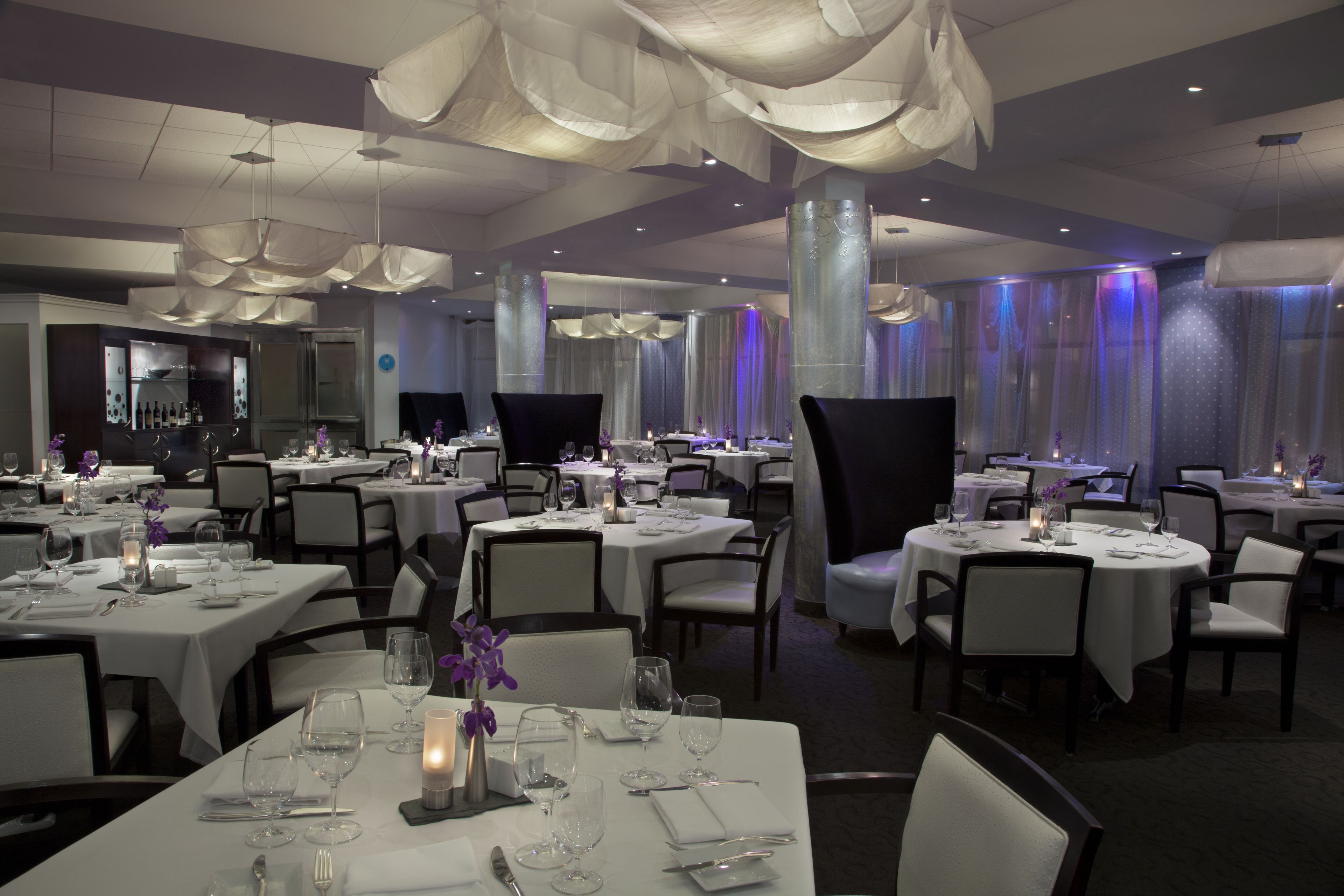 Un restaurante con varias mesas redondas, manteles blancos y candelabros modernos.