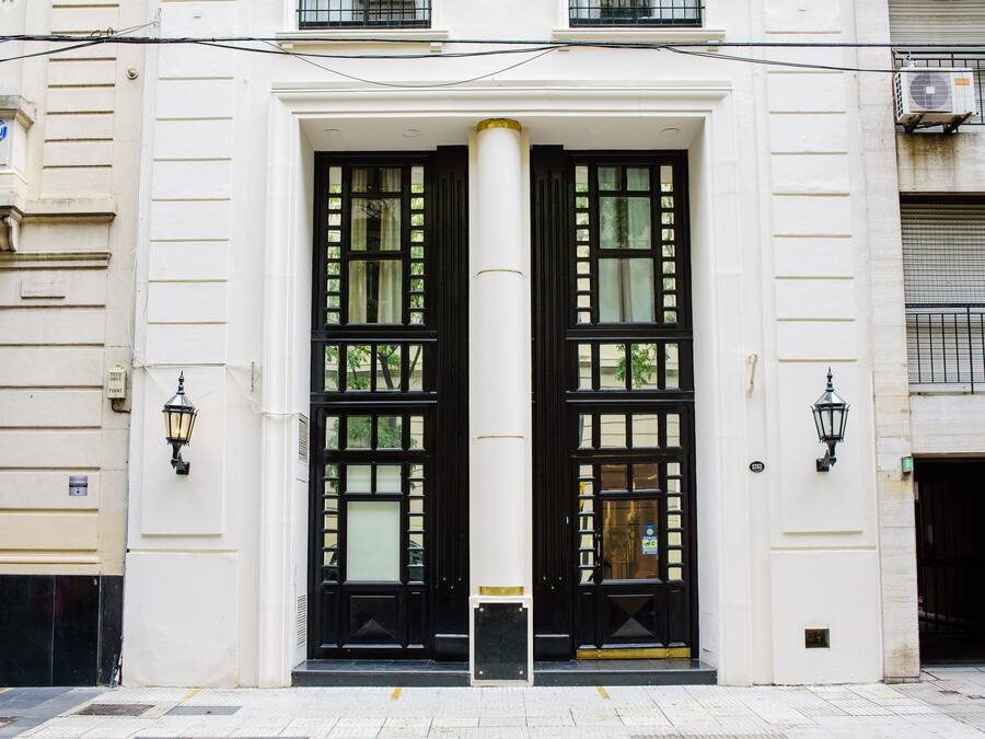 Fachada de edifício com entrada central destacada por duas grandes portas pretas com painéis de vidro, ladeadas por duas luminárias pretas em estilo lanterna e um pilar branco ao centro.