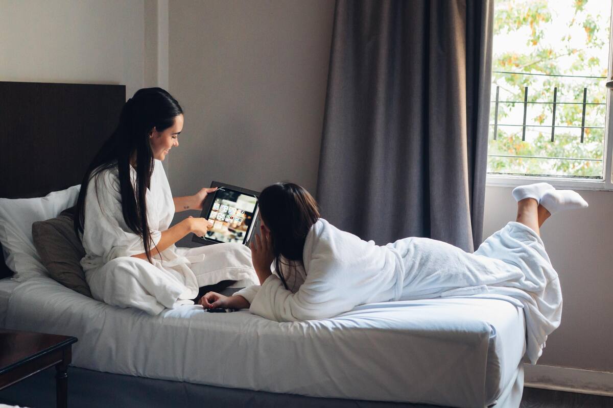 A room with a bed, a window with a view of greenery, and a person holding a tablet.