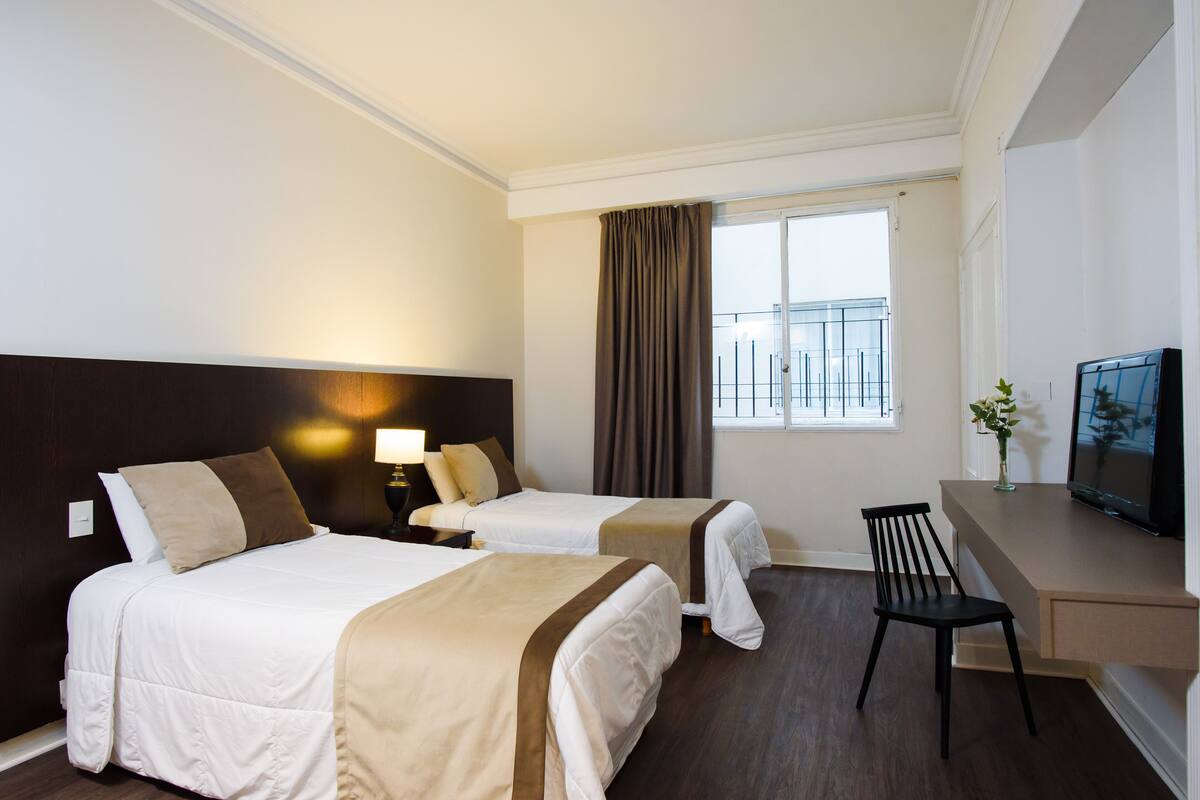 A hotel room with two beds, a desk with a chair, a TV, and a window with curtains.