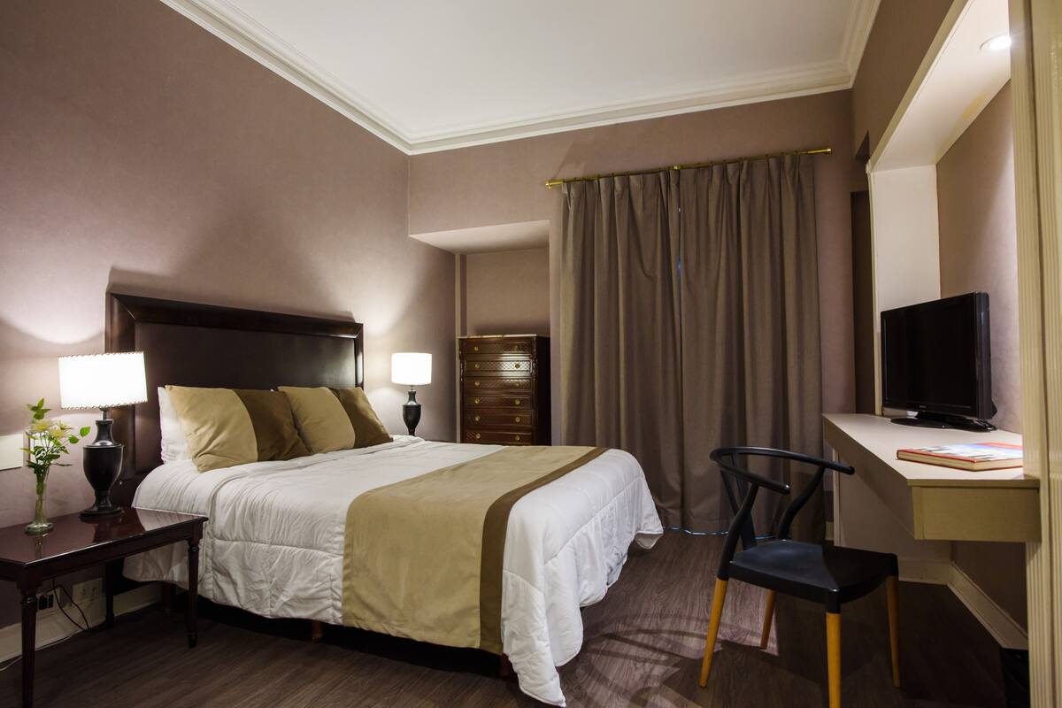 A hotel room with a bed, desk, chair, television, and a nightstand with a lamp and vase.