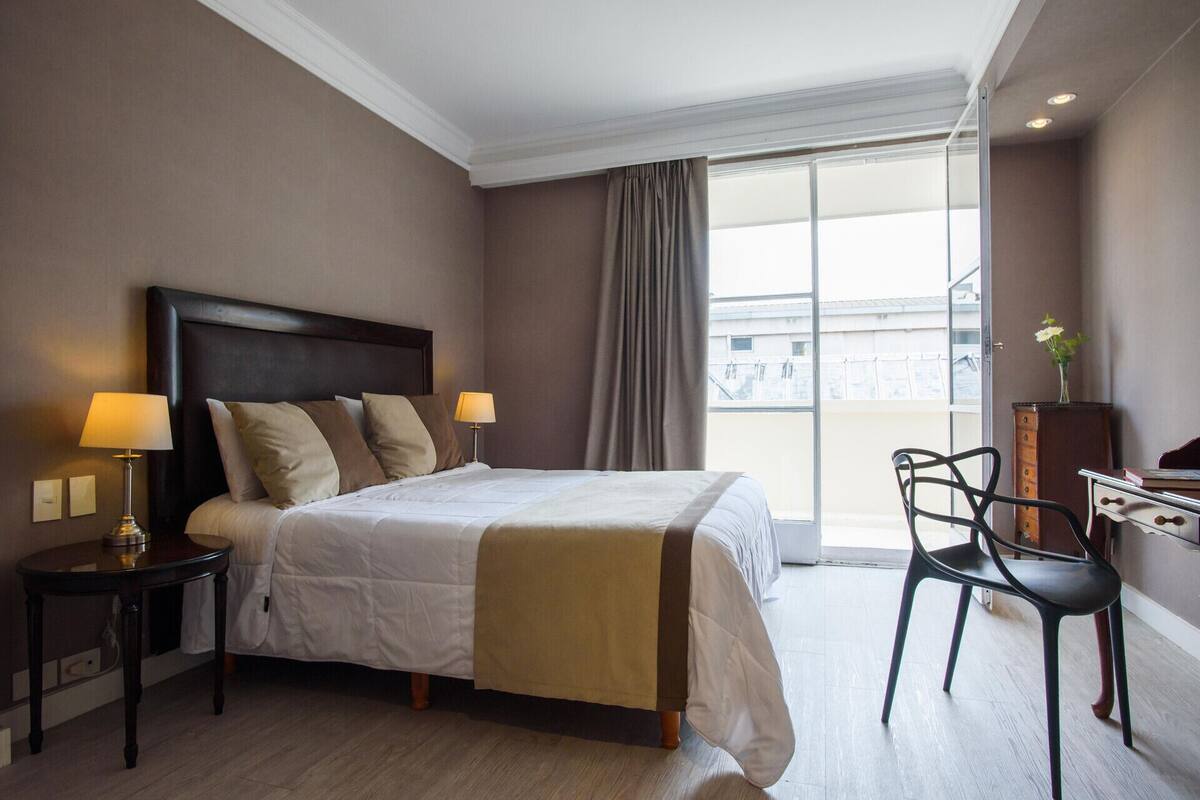 A hotel room with a large bed, two bedside lamps, a desk with a chair, and a view of buildings through a large window.