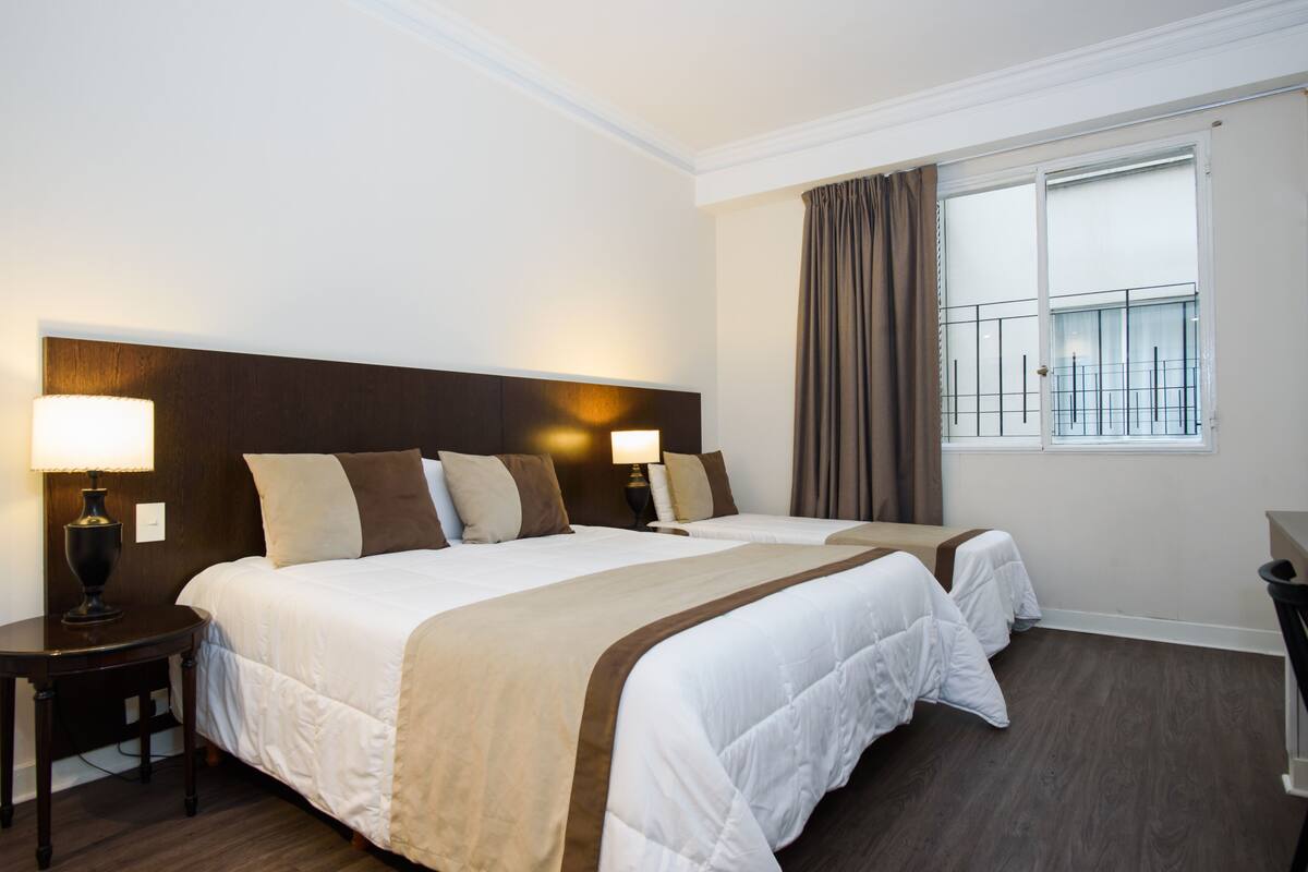 A hotel room with two beds, a large window with curtains, a nightstand with a lamp, and a view of the city skyline.