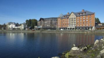 A large hotel with multiple floors and balconies, situated by a calm lake with rocky shoreline.