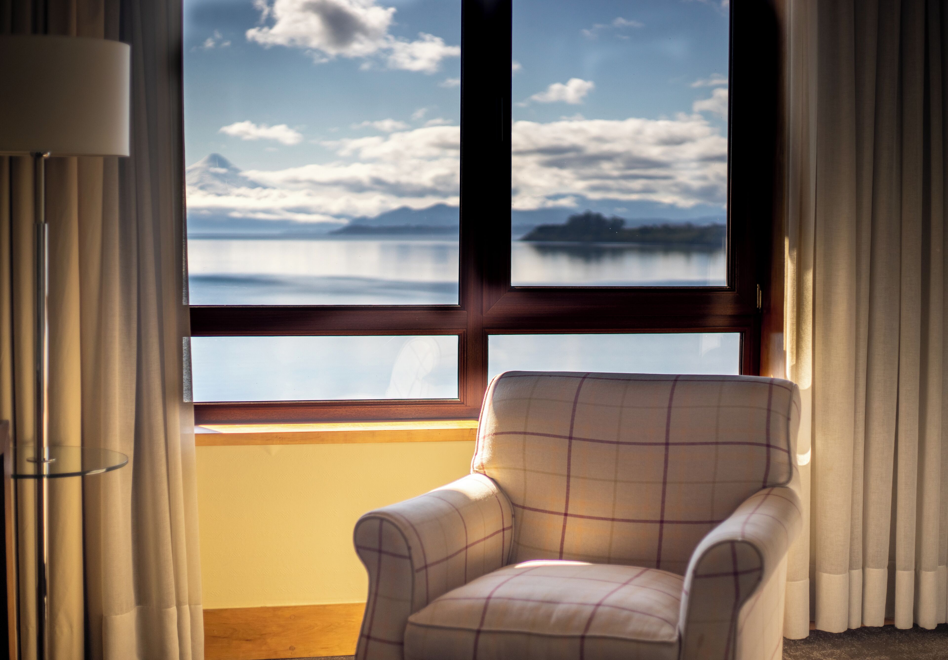 A room with a view of a lake and mountains through a window, a chair, and curtains.