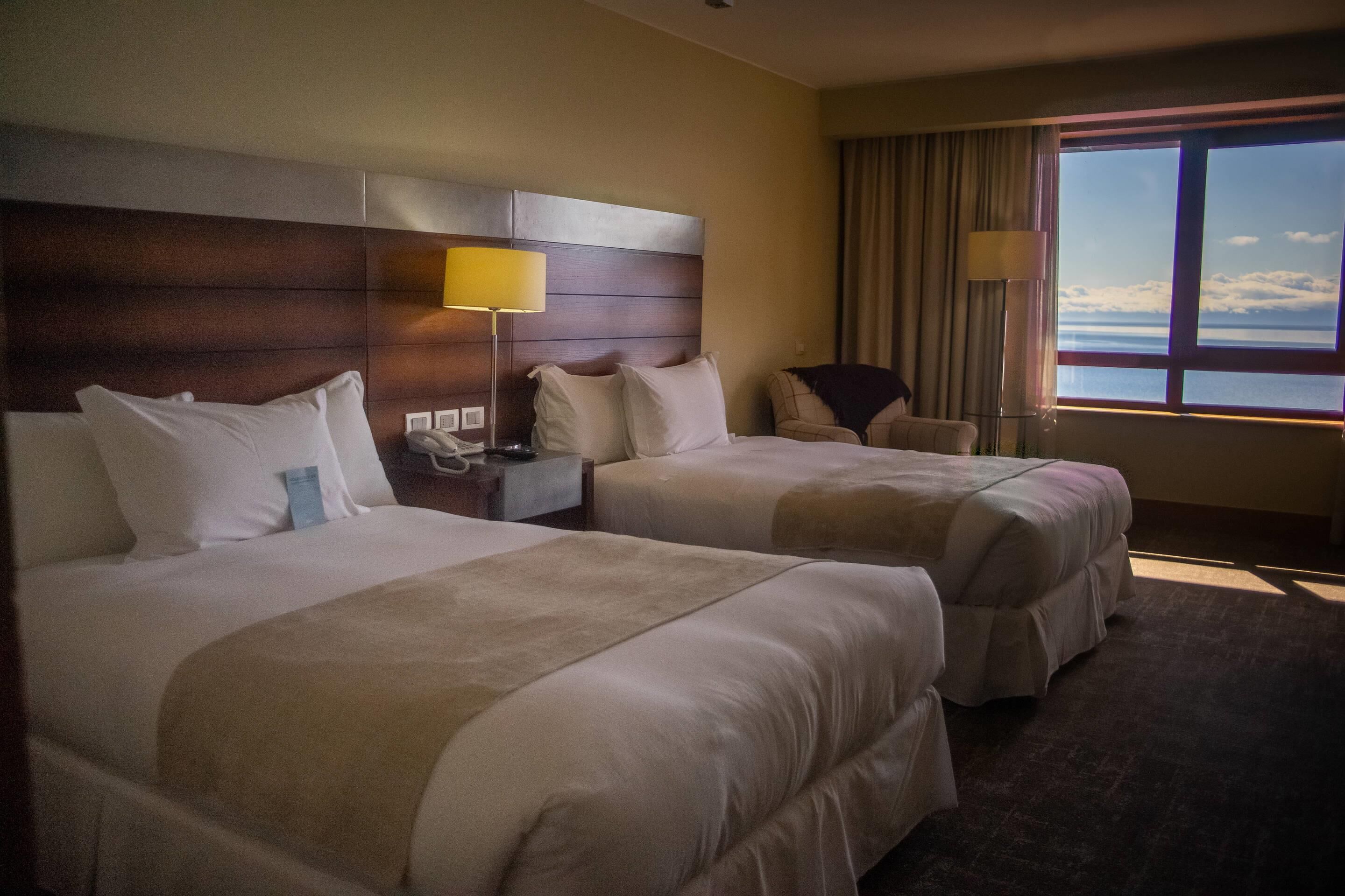 Une chambre d’hôtel avec deux lits, une table de chevet, une lampe, une chaise et une grande fenêtre donnant sur la mer.