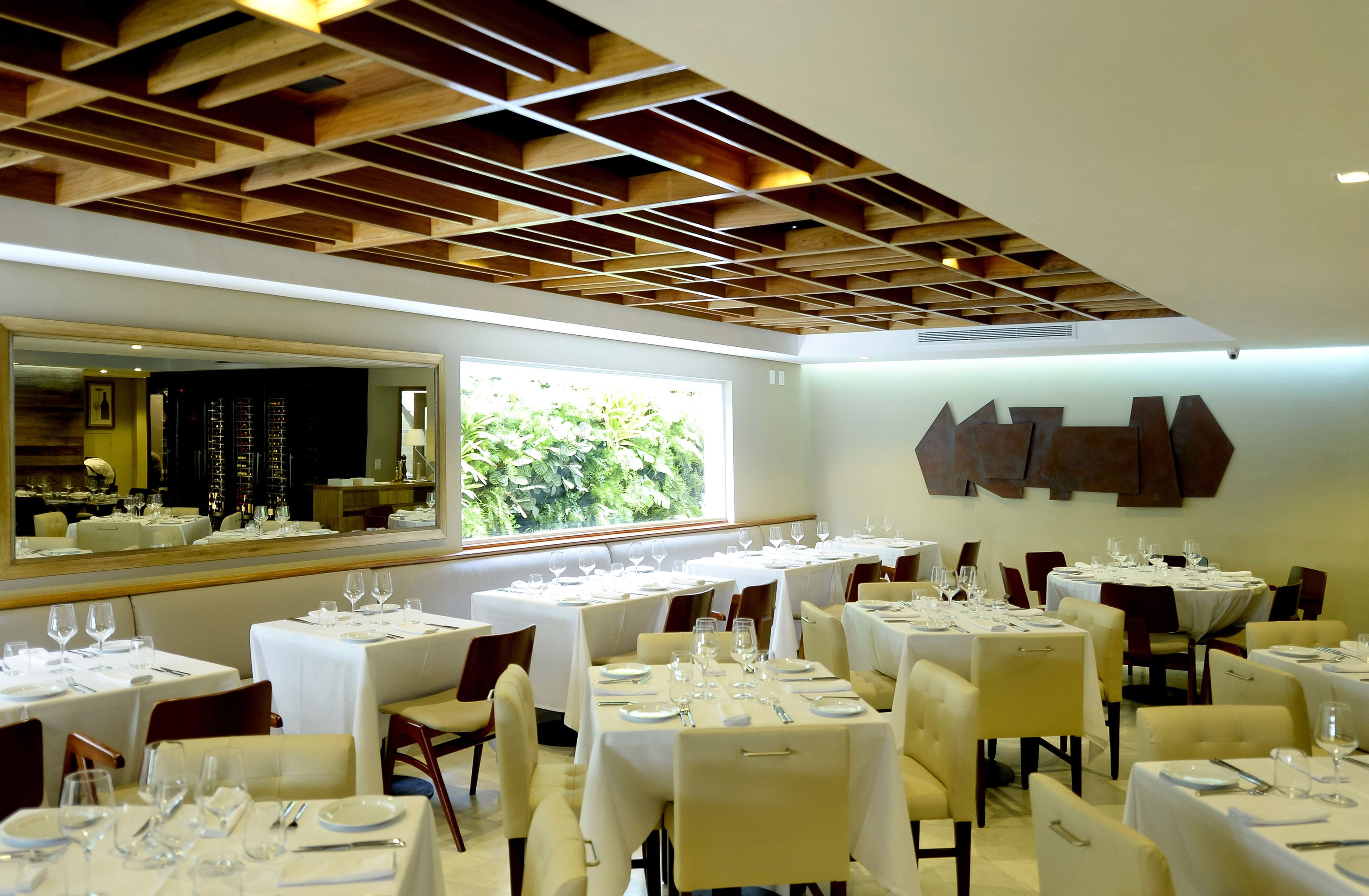 A restaurant with multiple tables set for dining, a large mirror, and a view of greenery outside.